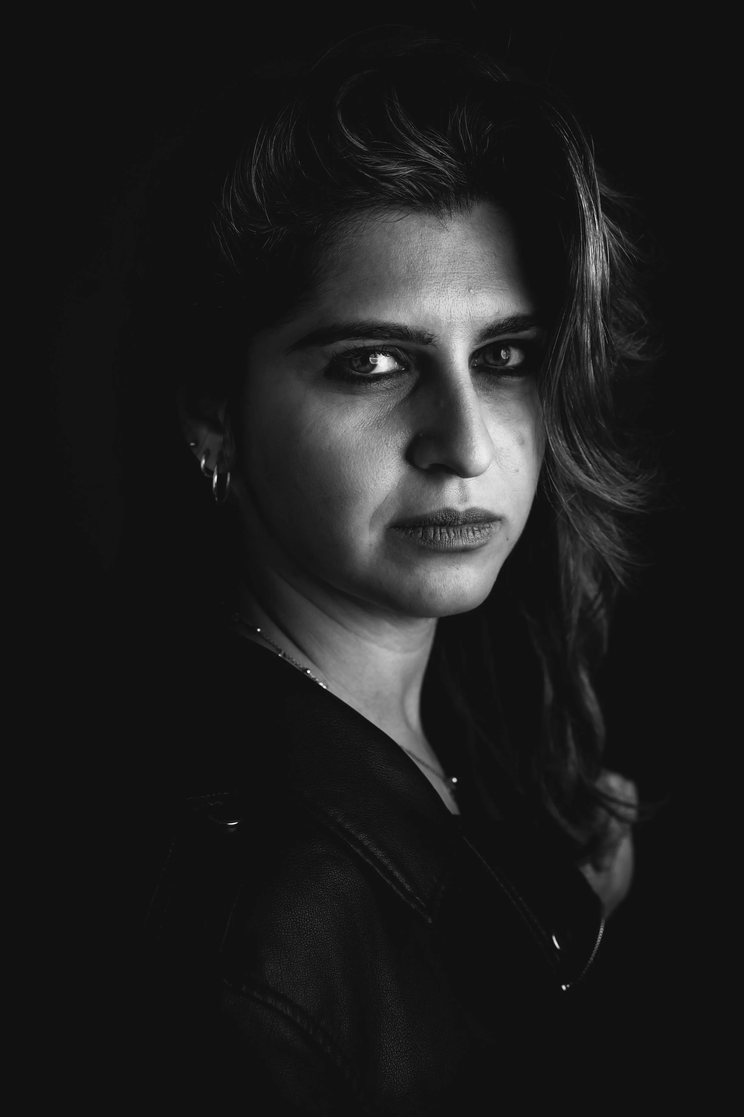 Ritratto in bianco e nero di una donna con capelli lunghi e ondulati, indossa orecchini a cerchio e una giacca, con uno sguardo diretto verso l'obiettivo, sfondo nero.