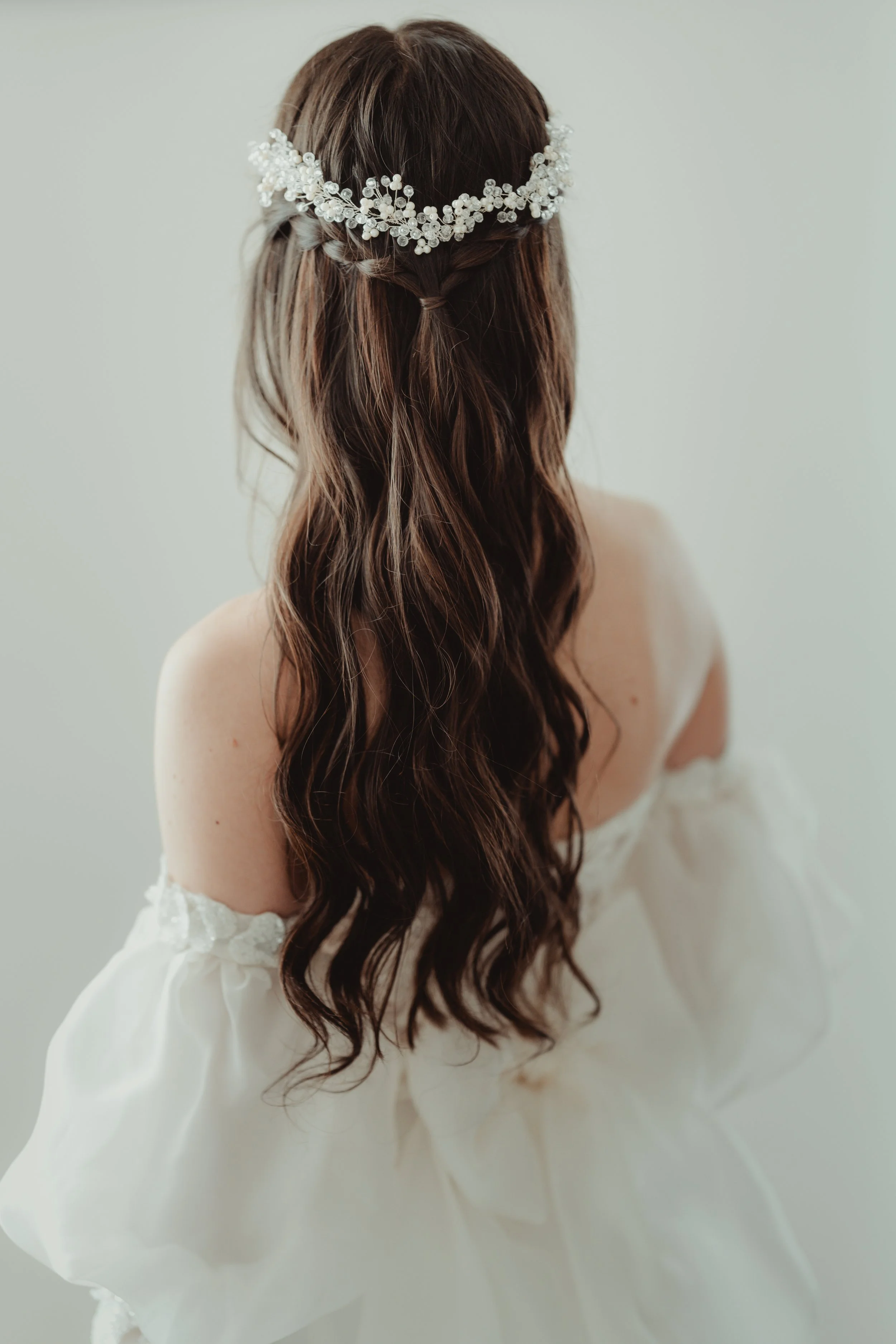 Capelli lunghi e mossi con corona di fiori, abito bianco, sfondo neutro.