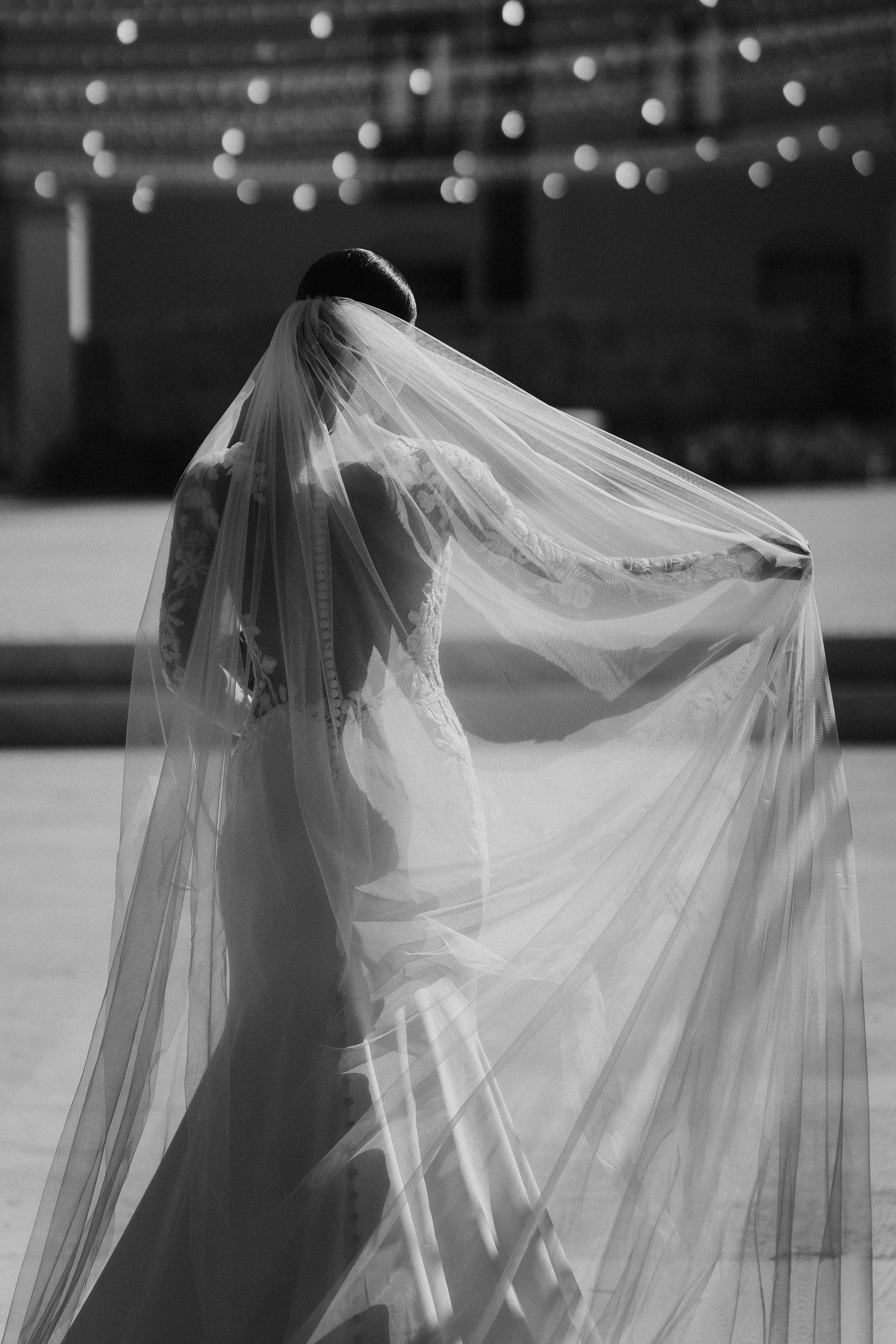 Sposo con abito da matrimonio e velo che si tiene la gonna mentre cammina all'aperto, sfondo sfocato con luci decorative.