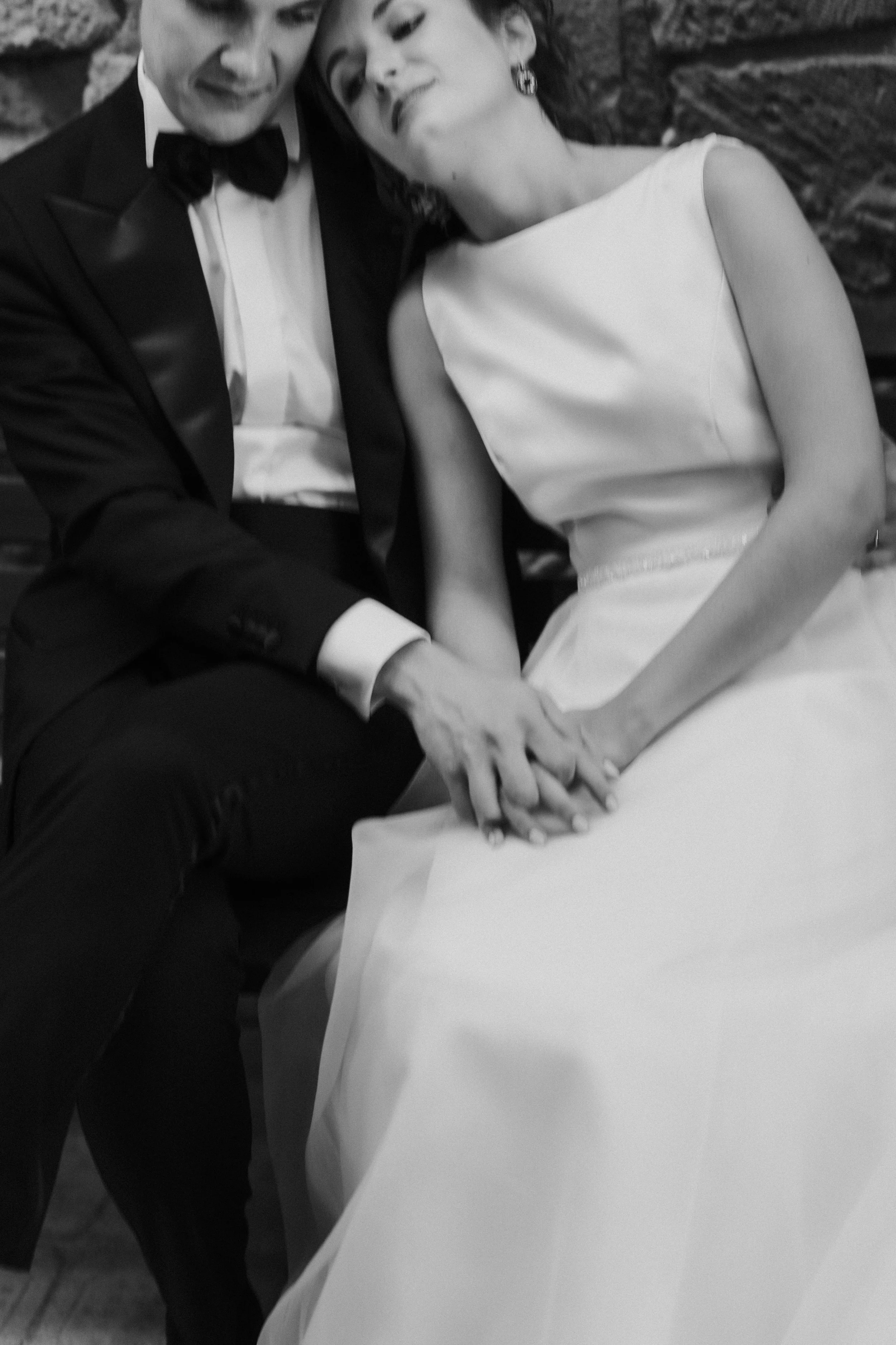Due persone, un uomo in abito formale e una donna in abito bianco, si tengono per mano e sembrano essere felici, sullo sfondo una parete di pietra.