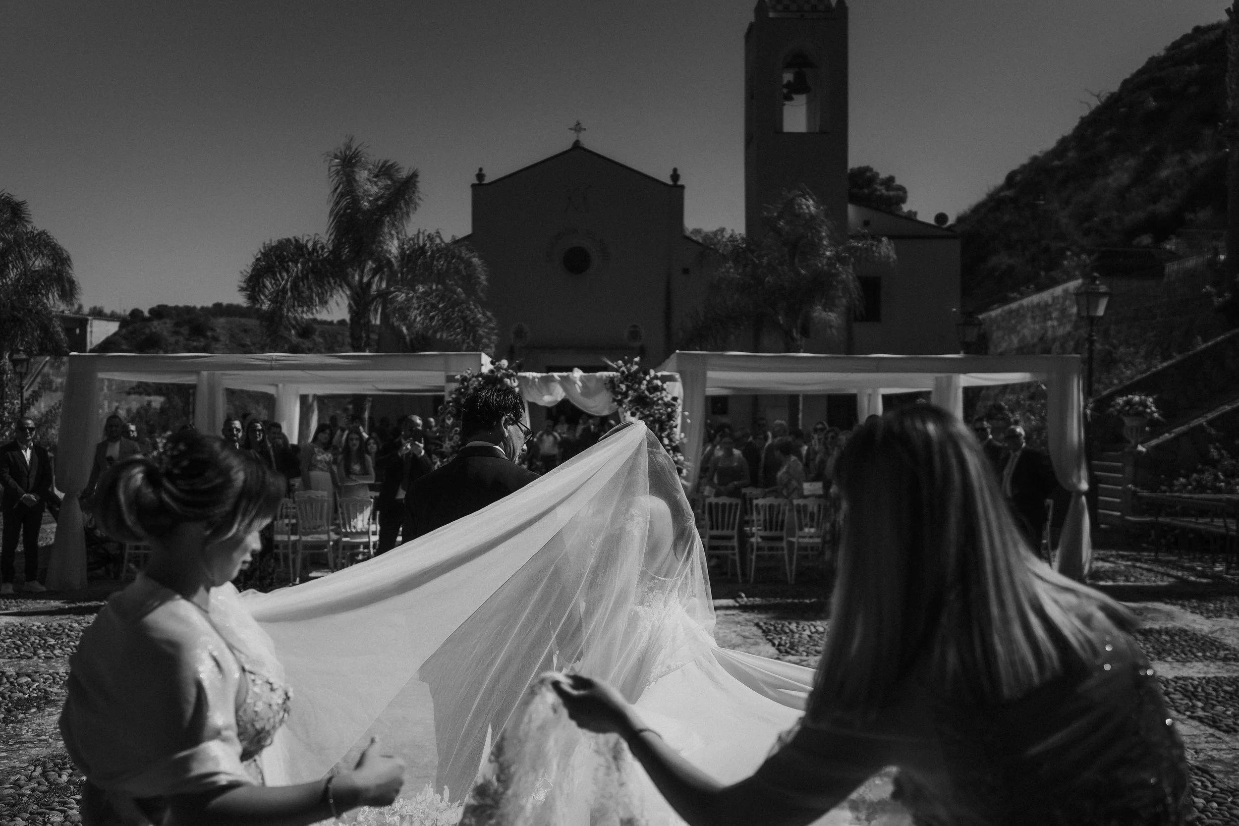 Una coppia di sposi si sposa all'aperto davanti a una chiesa con un gruppo di testimoni e amici, con montagne sullo sfondo e decorazioni floreali.
