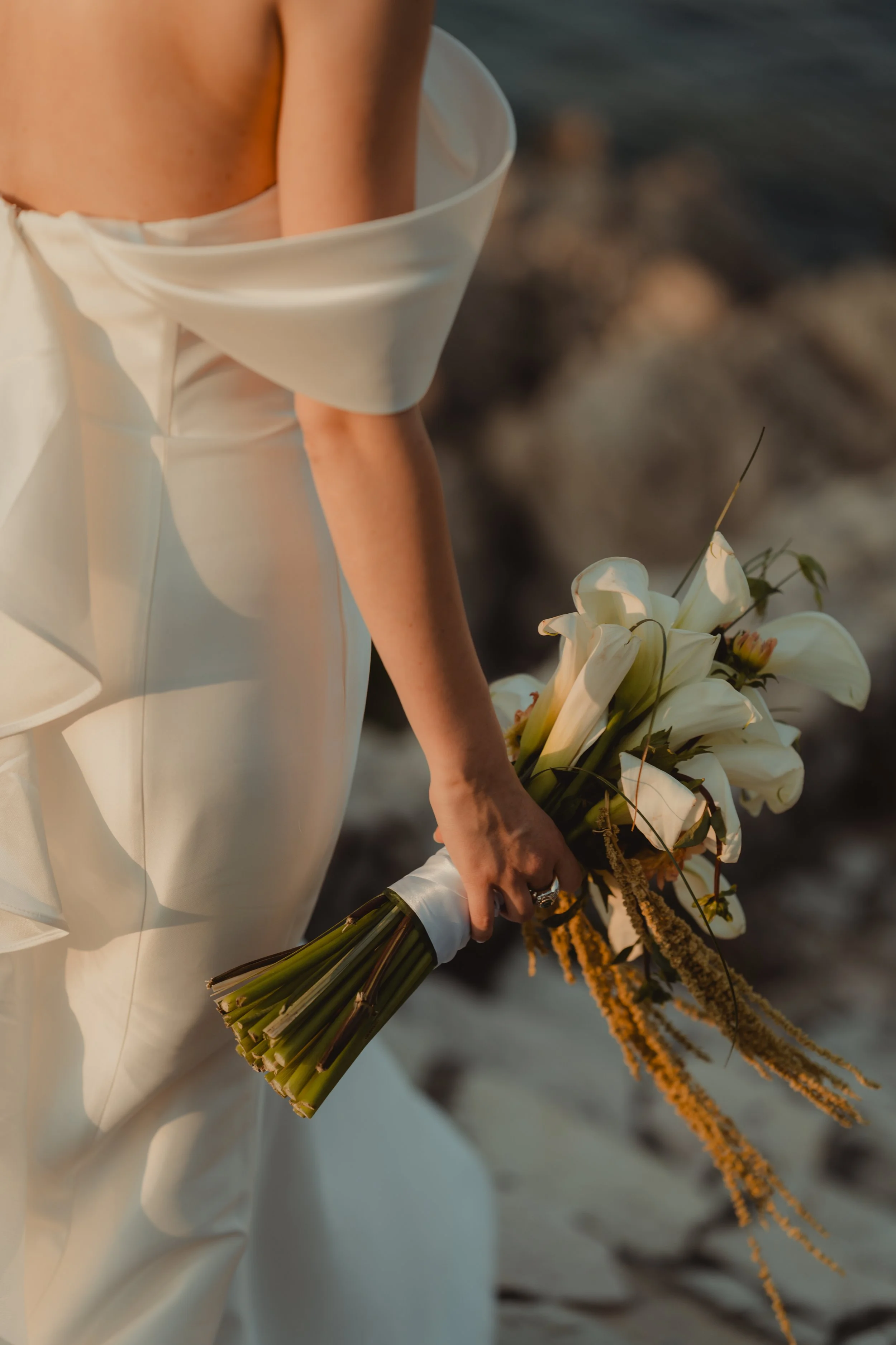 Donna in abito bianco stringato che tiene un mazzo di fiori bianchi e gialli, con uno sfondo naturale.