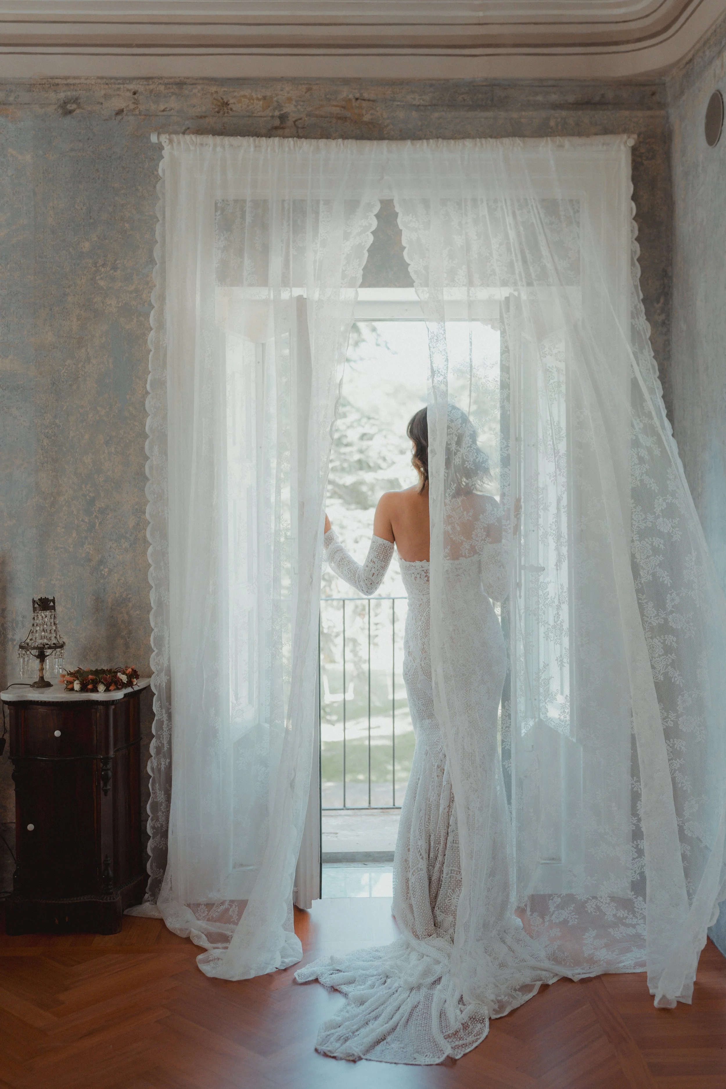 Donna in abito da sposa che guarda fuori una finestra con tende di pizzo bianco, in una stanza con pareti dipinte o decorate.