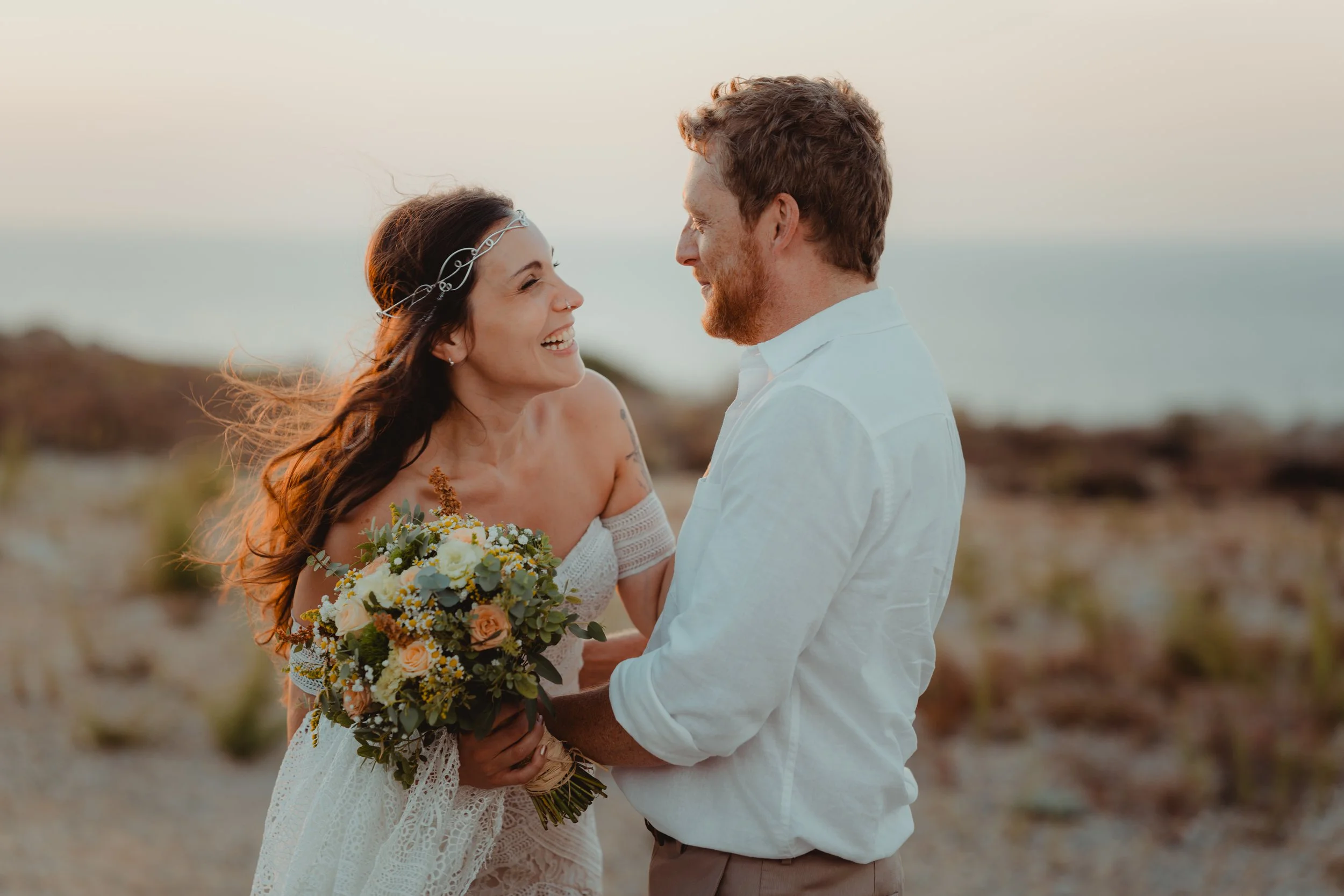 Una coppia felice si guarda negli occhi sulla spiaggia al tramonto, con la donna che tiene un mazzo di fiori. Sono sorridenti e innamorati.