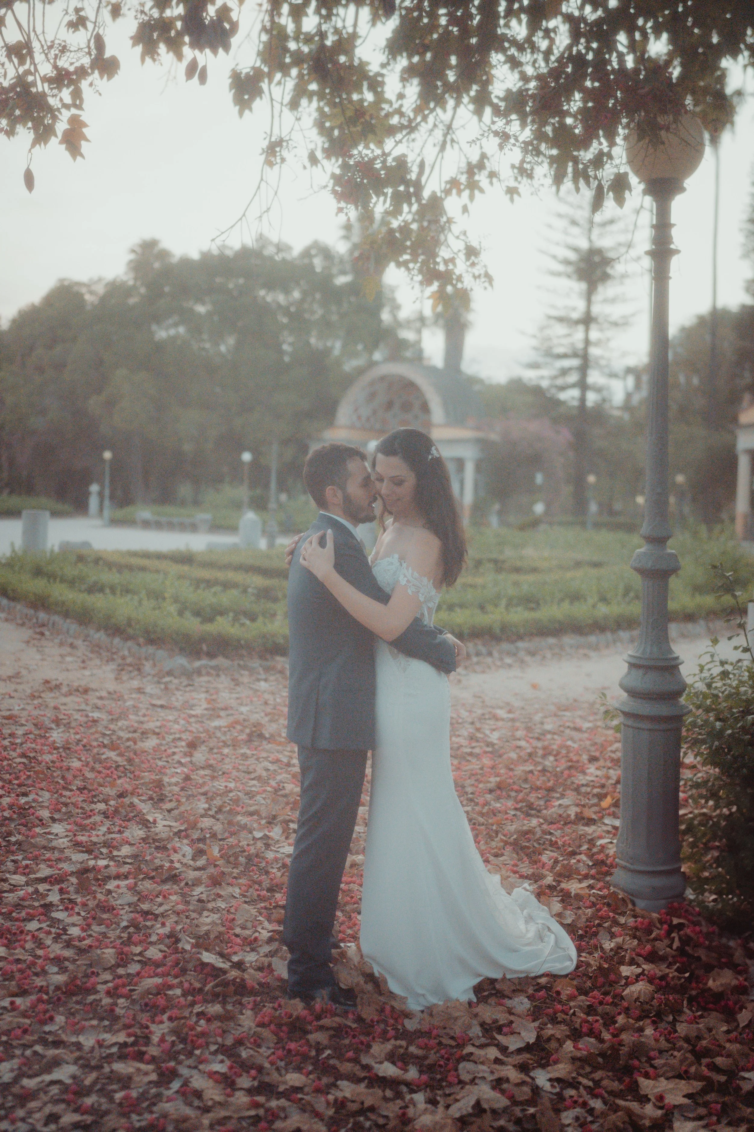 Una coppia in abiti da matrimonio si abbraccia e si guarda negli occhi, con una cornice di alberi e foglie autunnali sullo sfondo in un parco. Una lanterna in stile vittoriano si trova sul lato destro.