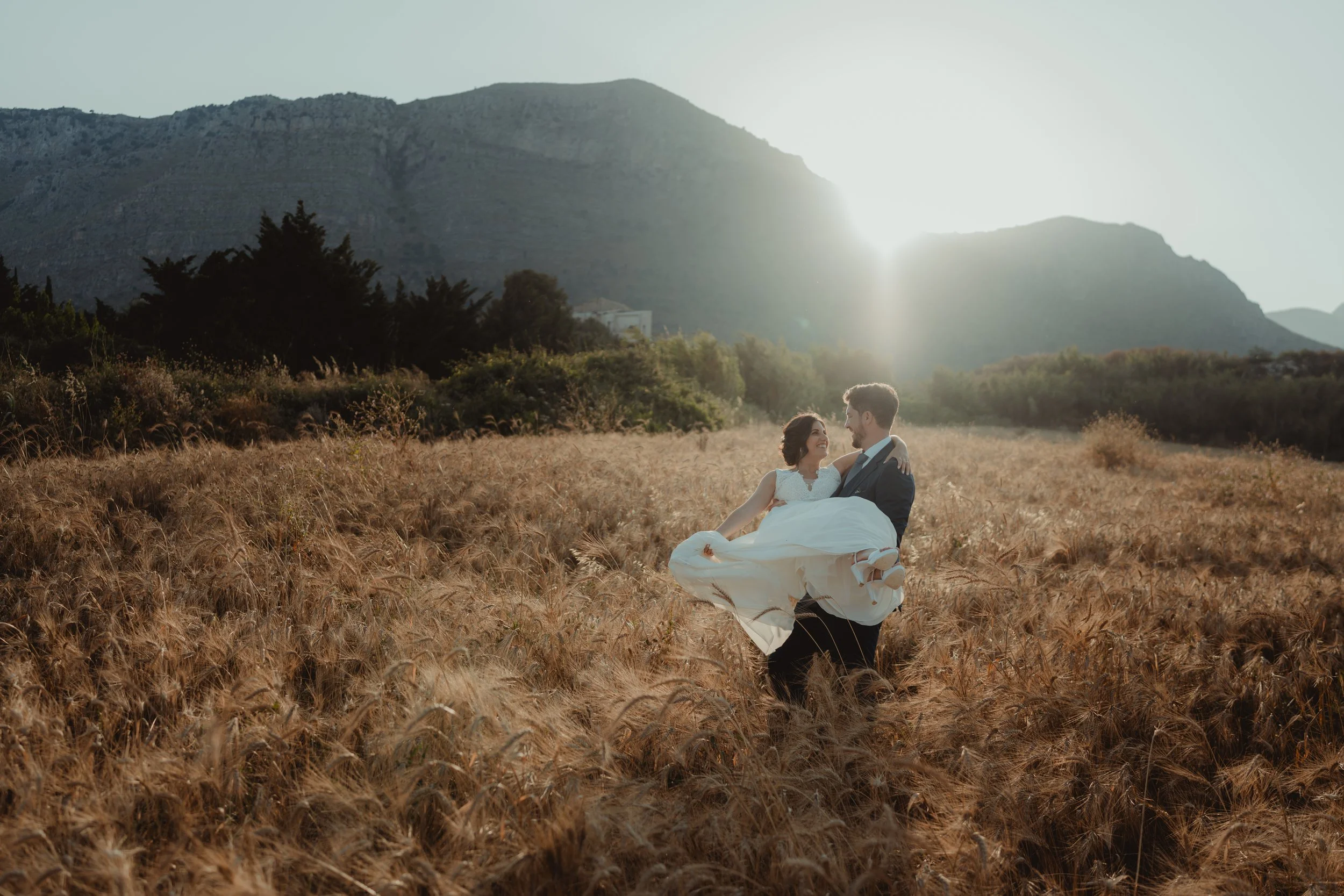 Una coppia in abiti da matrimonio si trova in un campo di grano dorato con montagne sullo sfondo e il sole che splende alle loro spalle.