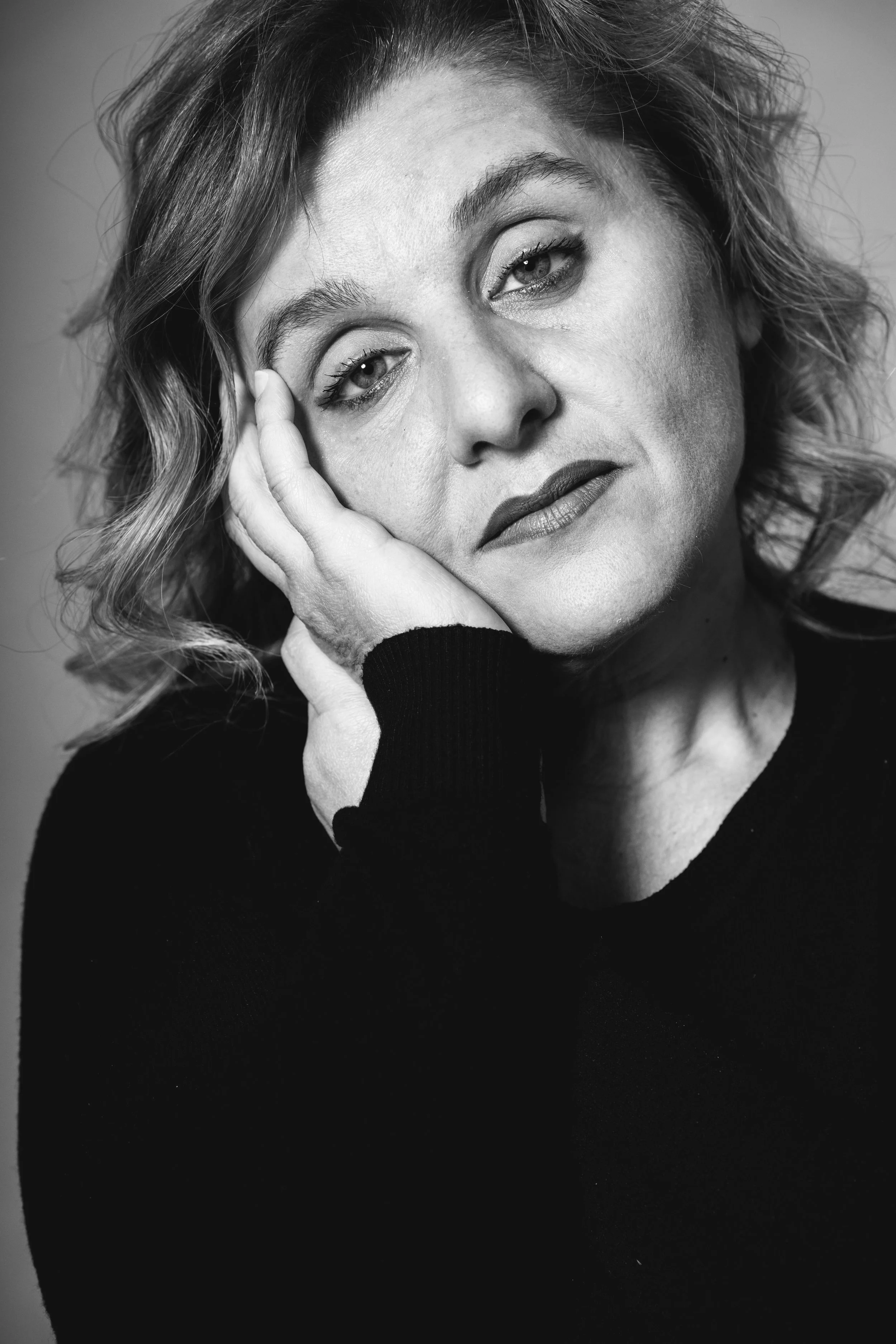 Ritratto in bianco e nero di una donna che sorride leggermente, poggiando una mano sulla fronte, con capelli ricci e occhi luminosi.