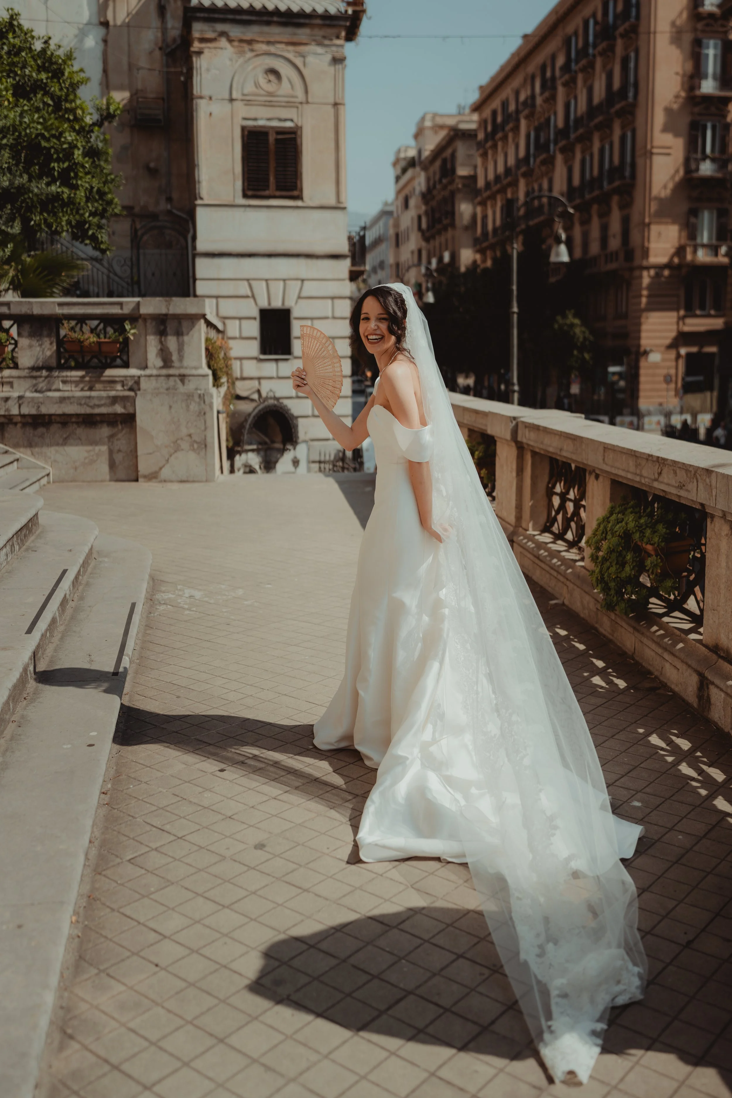 Una sposa sorridente in abito bianco con velo, che tiene un ventaglio di legno, in piedi su una scalinata di pietra in un ambiente urbano.