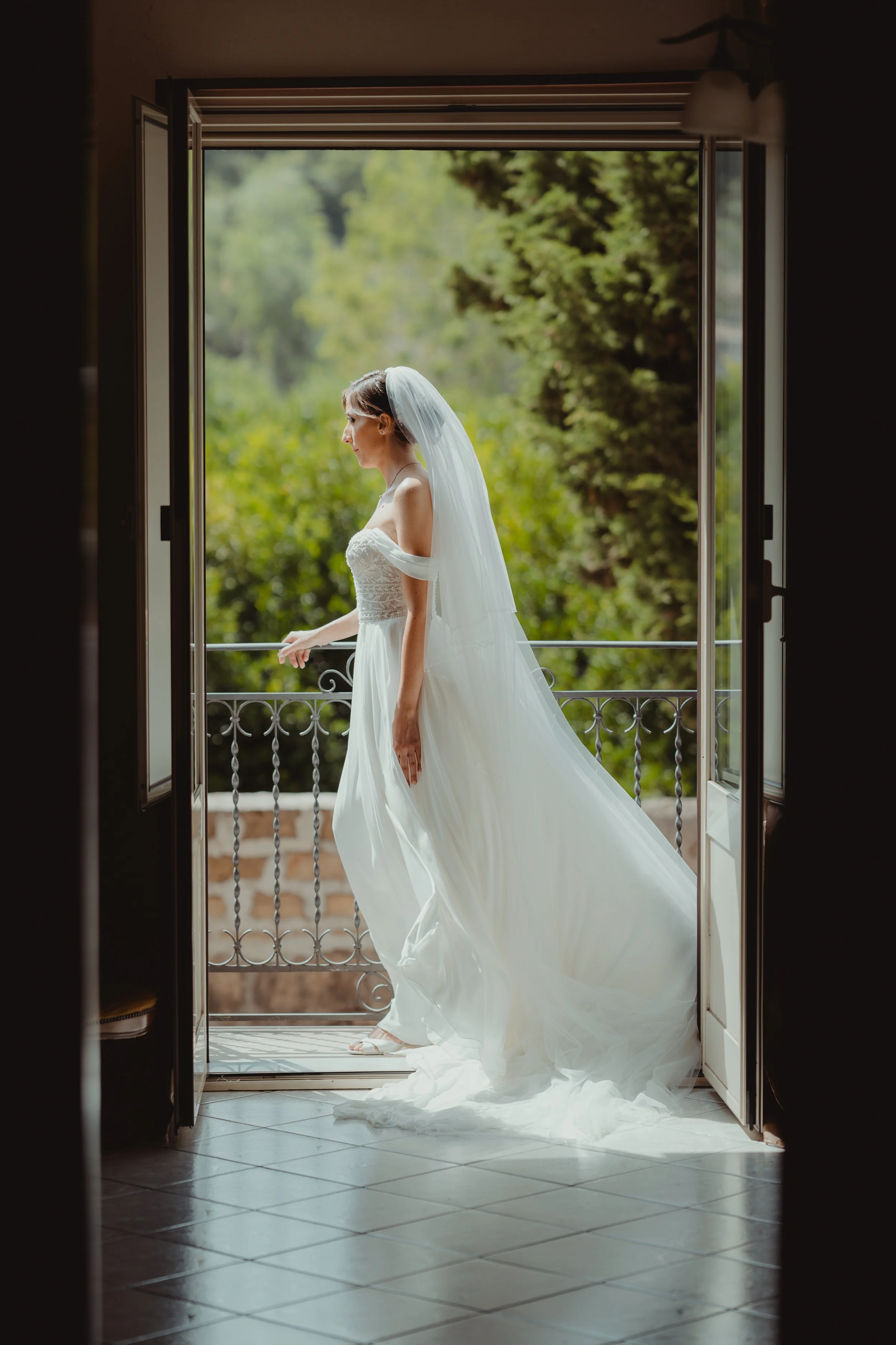 Una sposa in abito bianco con velo si trova su un balcone, vista da dentro casa in una stanza scura, con uno sguardo pensieroso verso l'esterno, circondata da natura verde.