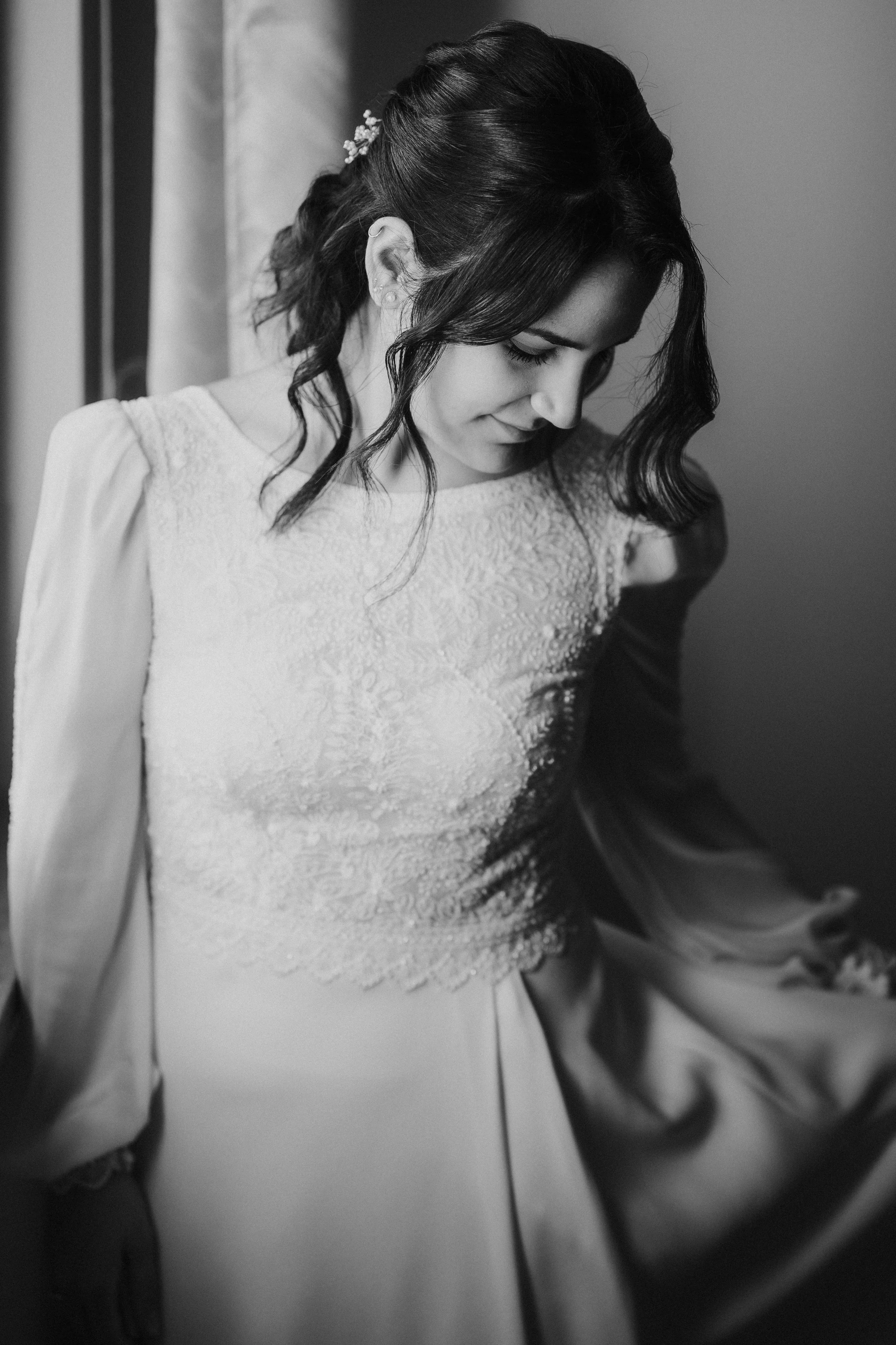 Ragazza con capelli scuri e riccioli, vestita in un vestito bianco, si guarda nello specchio con un sorriso delicato, sfondo sfocato con parete chiara.