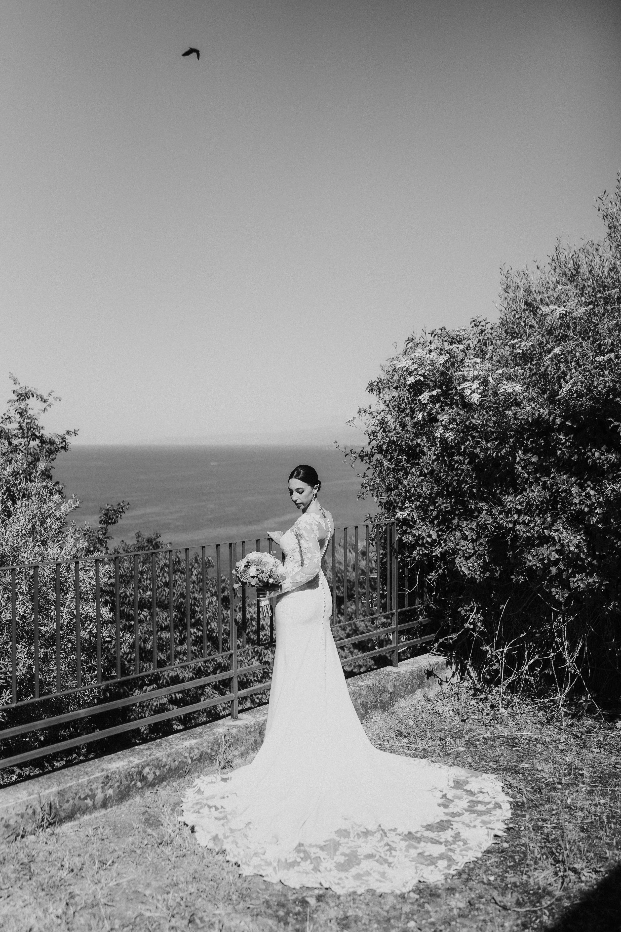 Una donna in abito da sposa si trova in esterna, davanti a un cancello di metallo e con un paesaggio marino sullo sfondo, circondata da alberi.