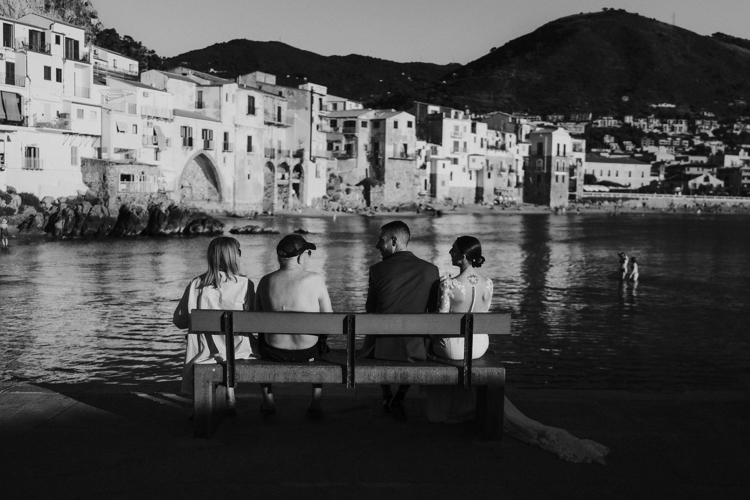 Quattro persone sedute su una panchina sulla riva del mare con un paesaggio di case e colline sullo sfondo