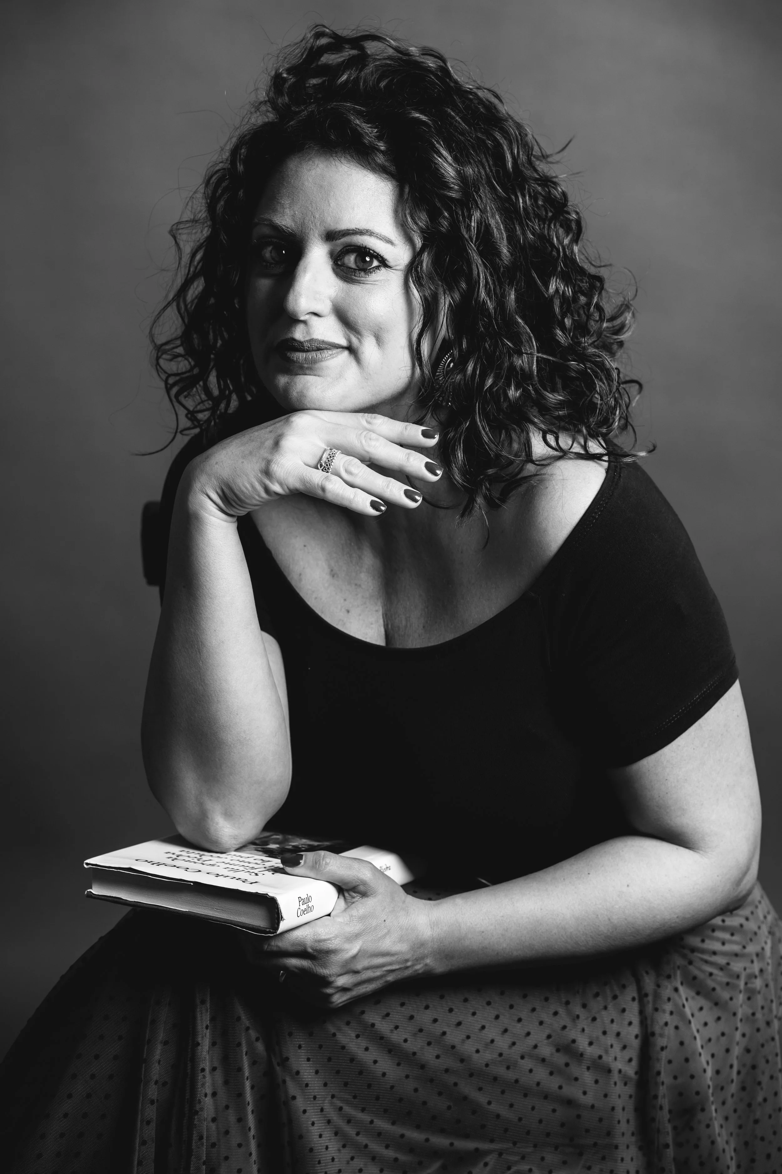 Una donna con capelli ricci e sguardo diretto, in posa frontale, tiene un libro sul grembo e poggia il mento sulla mano. È vestita con una maglietta nera e una gonna a pois. La foto è in bianco e nero.