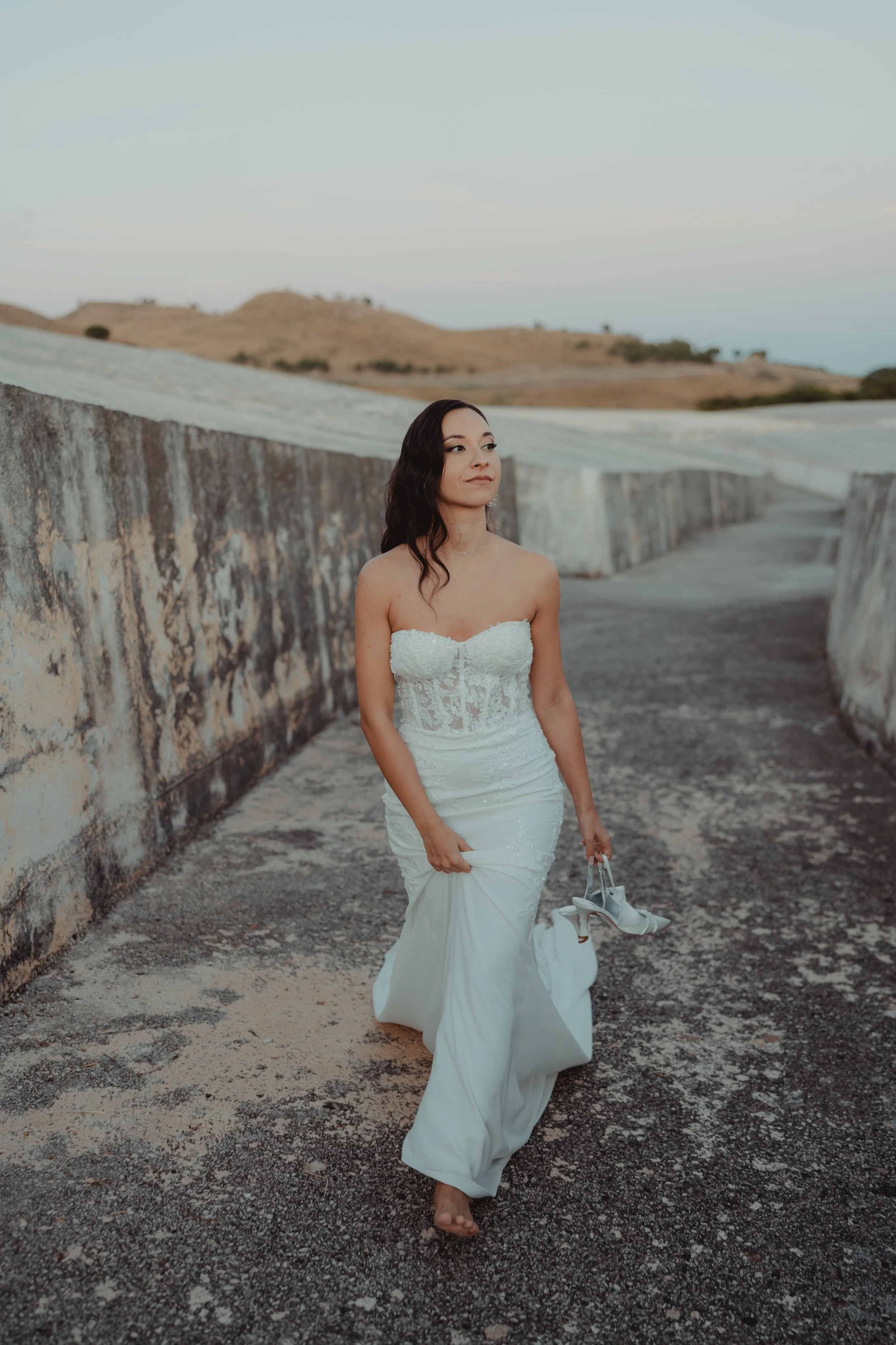 Una donna in abito da sposa cammina scalza lungo una passeggiata di pietra, tiene le scarpe in mano e guarda verso l'orizzonte con uno sfondo di colline e cielo azzurro.