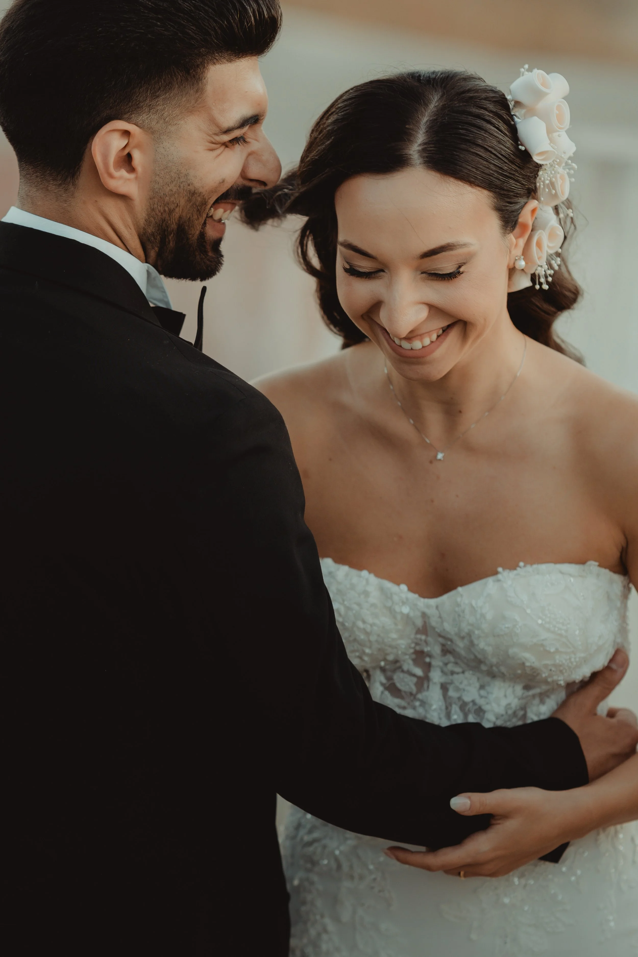 Una coppia di sposi sorridenti durante il loro matrimonio, con la donna che indossa un abito da sposa bianco e decorata con fiori in capelli, e l'uomo in abito scuro. Sono stretti insieme, quasi in un abbraccio.