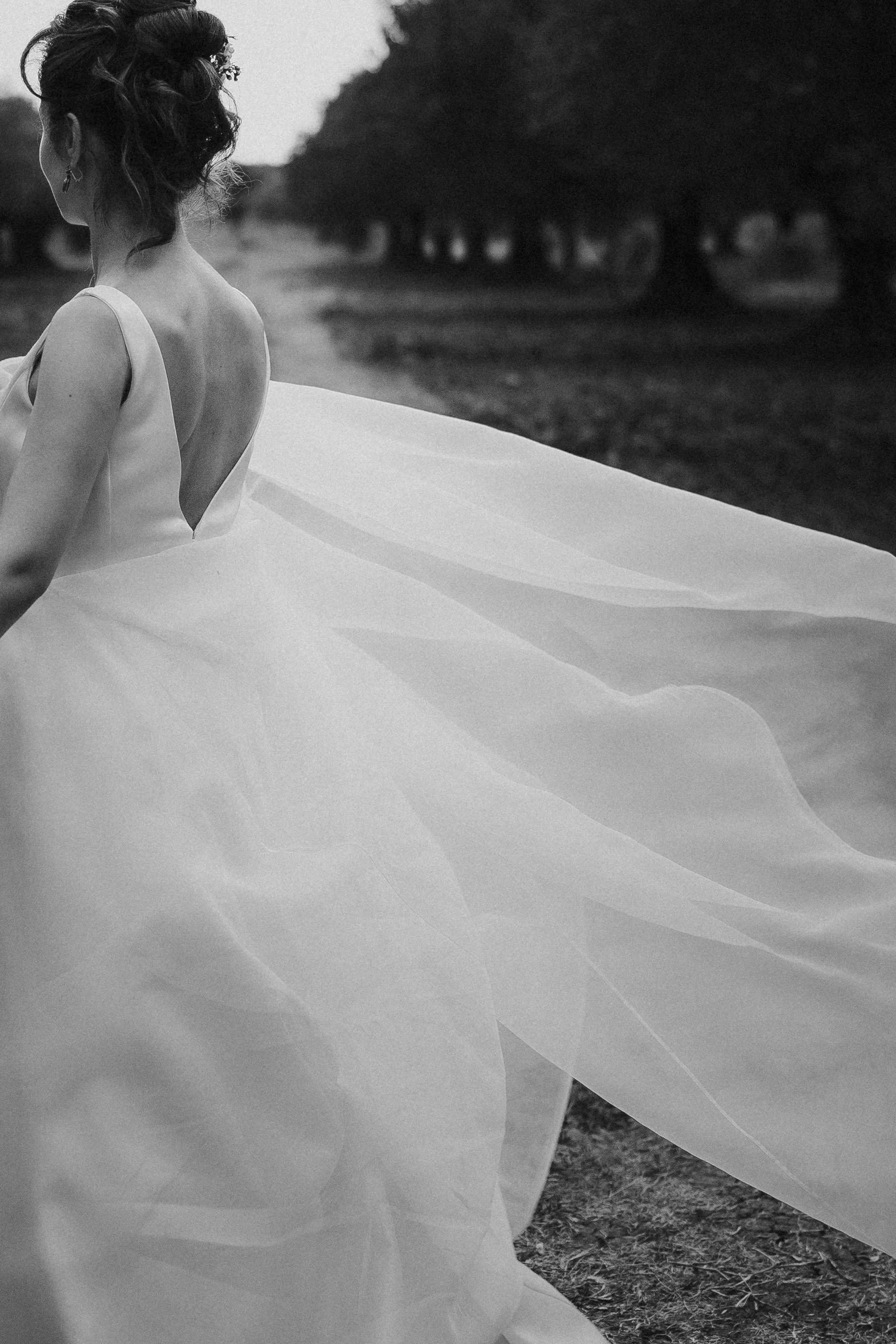 Donna con capelli raccolti, indossa un abito da sposa con schiena scoperta, che si muove al vento. Sullo sfondo un paesaggio naturale con alberi e terreno.
