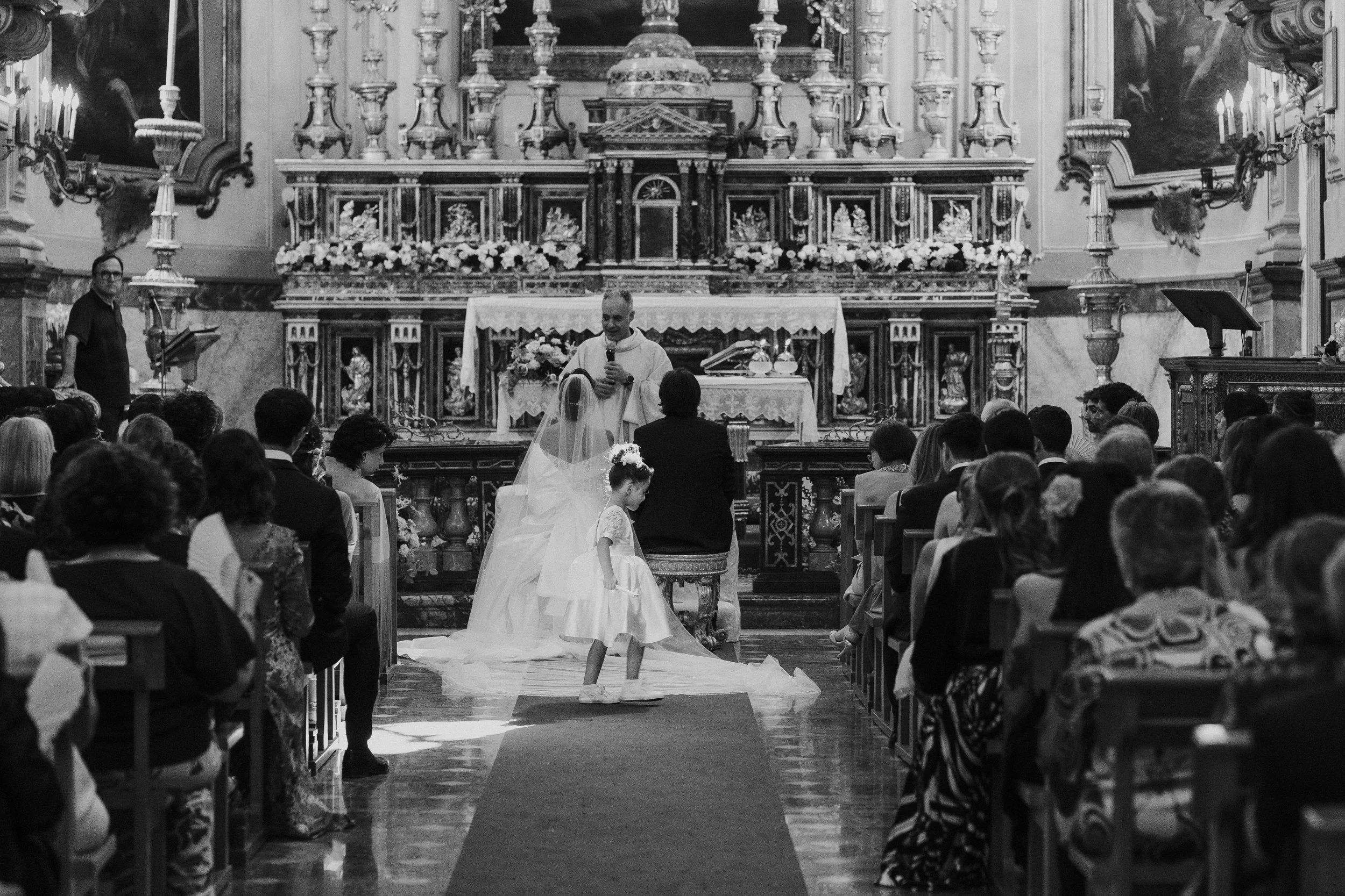 Un matrimonio in un' chiesa con molti ospiti seduti, una sposa con abito bianco e velo e un sacerdote che celebra il matrimonio di fronte a un altare decorato con fiori e statue.