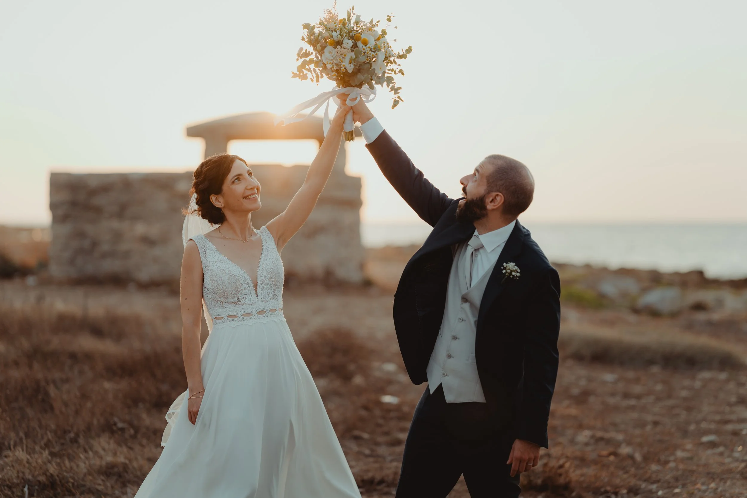 Una coppia di sposi si tengono per mano e si sorridono mentre la donna tiene un bouquet di fiori in aria, durante un tramonto all'aperto