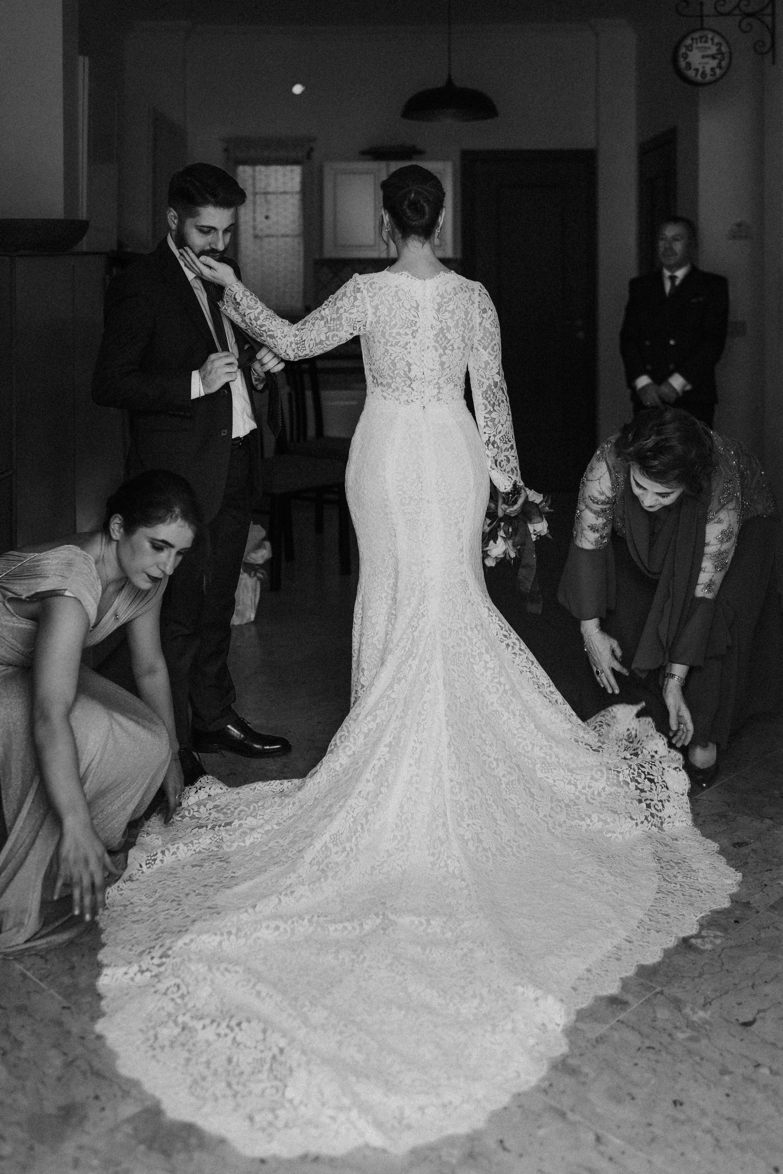 Una sposa in abito lungo di pizzo si sta sistemando prima del matrimonio, circondata da amiche e donne che la aiutano a sistemare il vestito. È in un ambiente interno, probabilmente una casa o una sala da ricevimento.