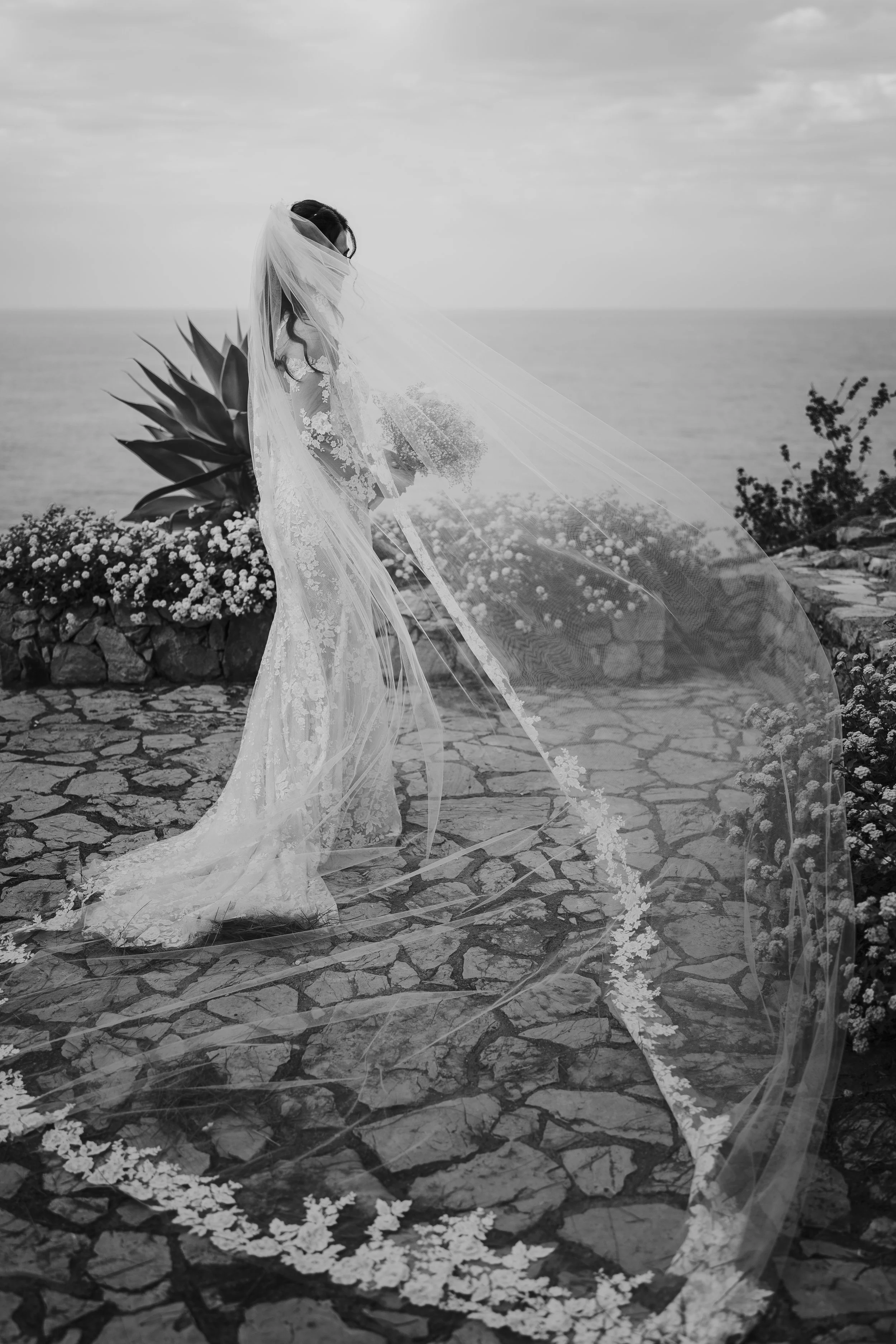 Una sposa con un abito lungo di pizzo e un velo trasparente si affaccia sulla strada di pietra, con il mare e il cielo nuvoloso sullo sfondo.