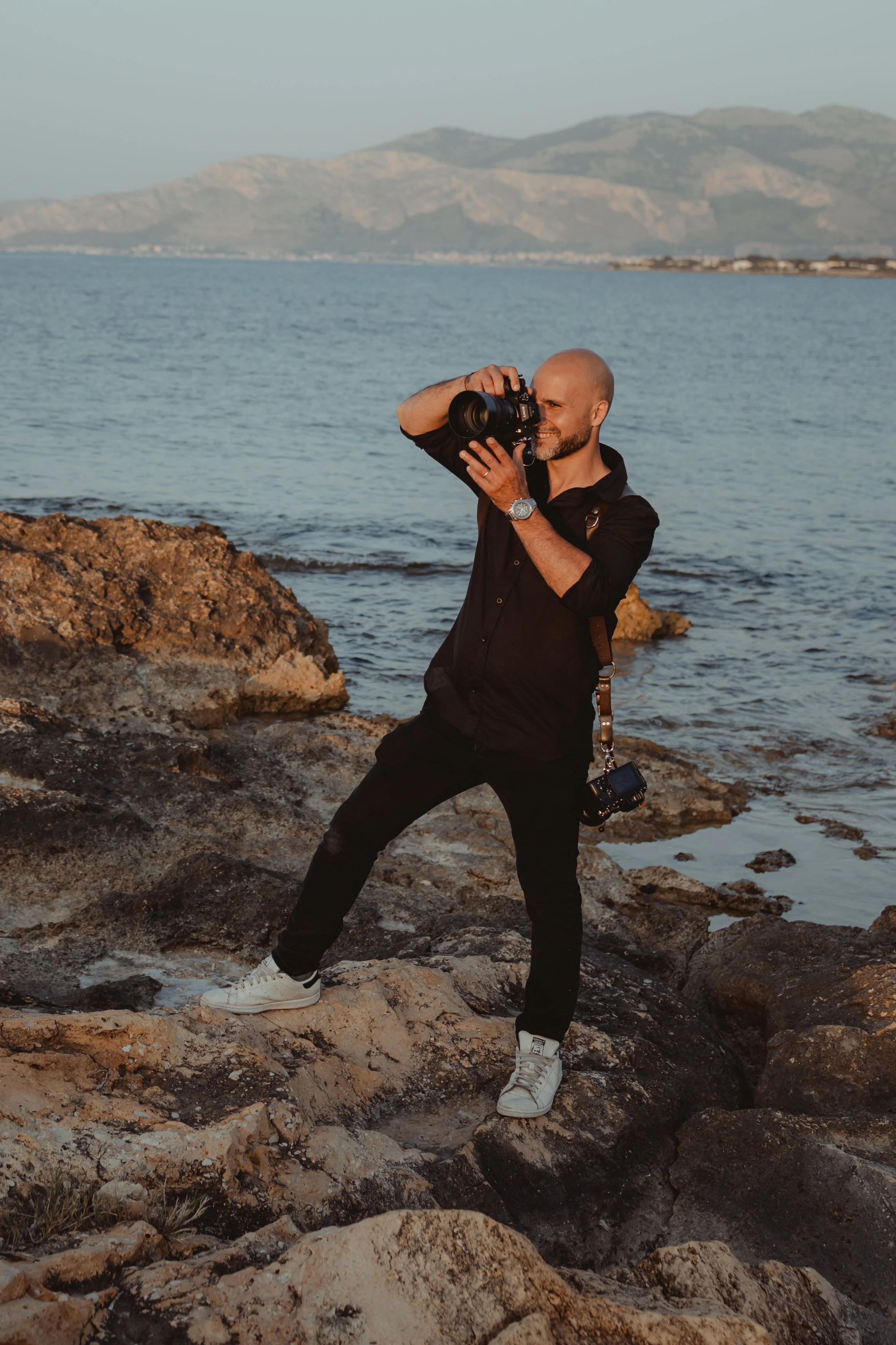 Vincenzo Latino che scatta fotografie sulla spiaggia con uno sfondo di mare e montagne all'orizzonte.