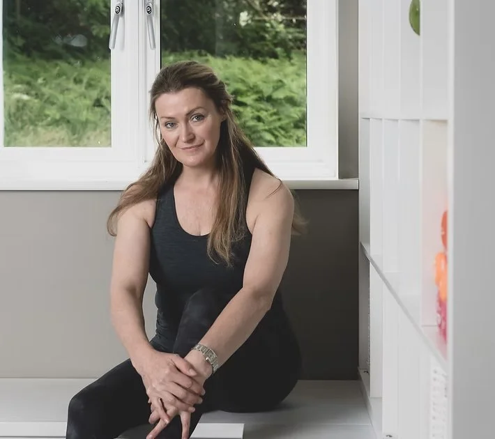 A woman sitting on a white bench by a window with greenery outside, wearing a black sleeveless top and pants, with brown hair and a watch on her left wrist.