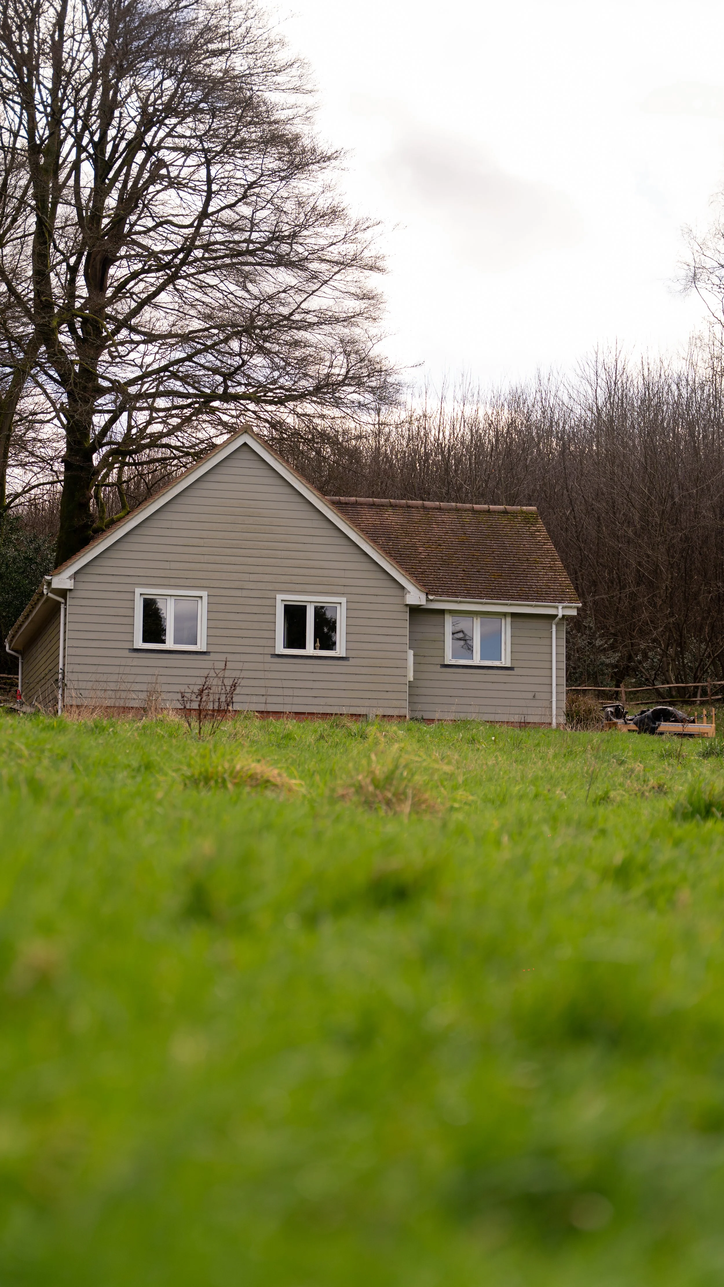 A gray house with white trim and a sloped roof, surrounded by a grassy yard and leafless trees in the background against a cloudy sky.