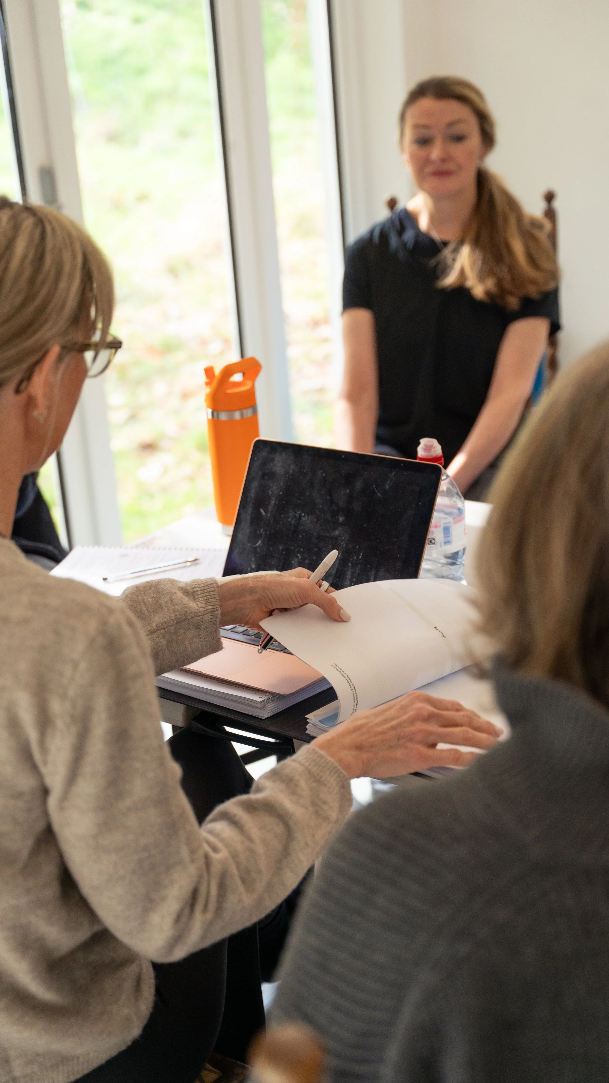 People gathered around a table in a bright room with large windows, discussing documents and using a tablet.