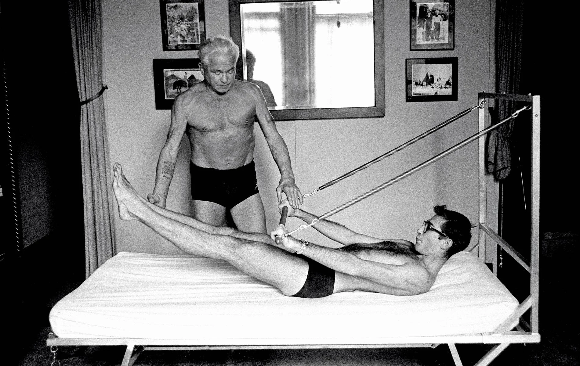 A black and white photograph of an elderly man with long hair lying on a bed, performing a leg stretch exercise with the aid of resistance straps, while a woman in glasses offers assistance and support. The room has framed pictures on the wall and a mirror reflecting light behind them.