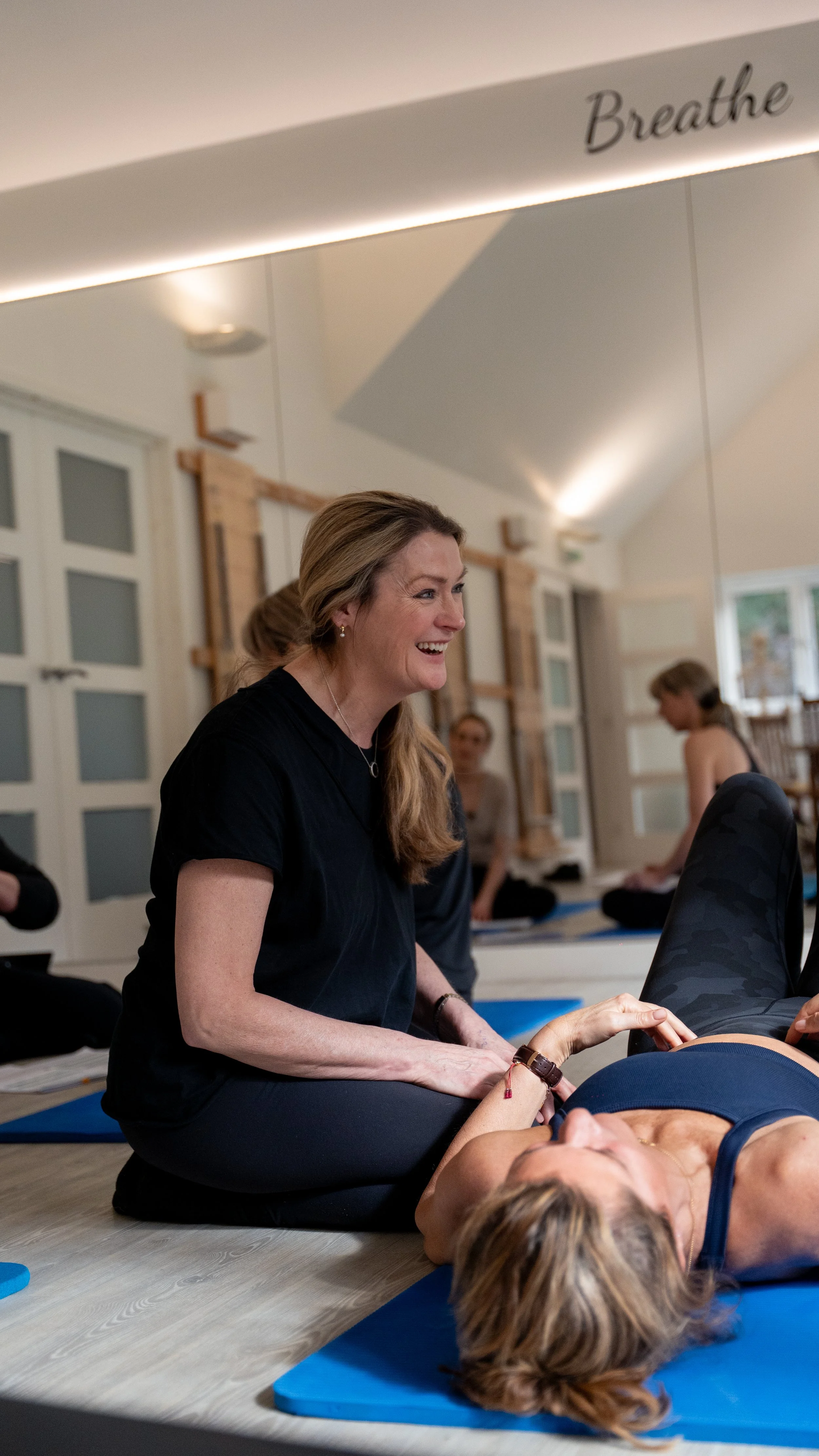 A woman in black clothing practicing yoga or meditation with a person lying on a yoga mat, while another woman helps or guides her, in a well-lit studio with large windows and wooden accents.
