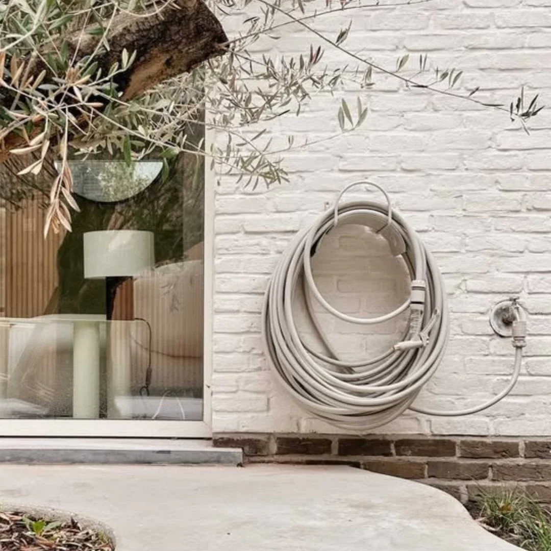 An outdoor scene showing a white brick wall with a garden hose coiled and hanging on it. There is a window to the left revealing a lamp and some furniture inside the house. A small patch of ground with some plants is seen at the bottom right corner.