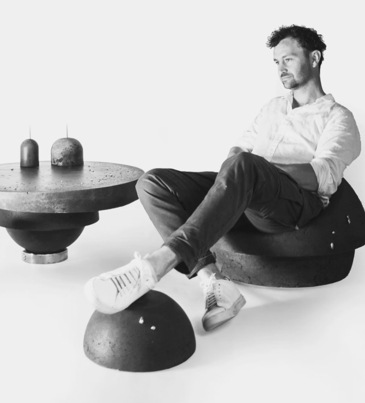 A man sitting on a modern, bean-shaped chair next to a round, textured table with two candles on it, in a minimalist setting.