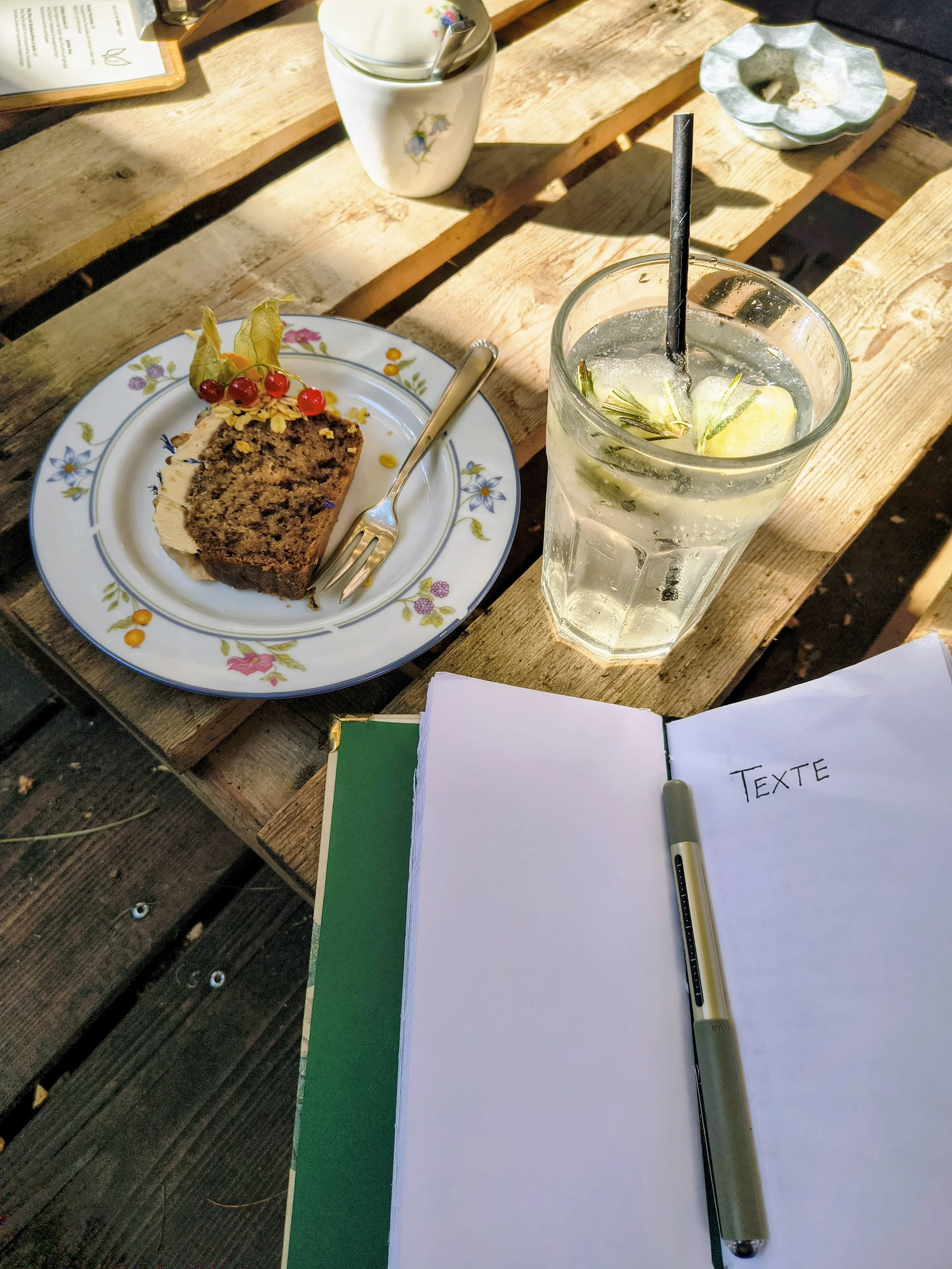 Ein Holzuntergrund mit einem gedeckten Tisch. Auf dem Tisch befindet sich ein Teller mit Kuchen, einer Gabel, einer Tasse mit Blumenmuster und einem Löffel, eine Glas mit Wasser, Zitrone, Rosmarin und Strohhalm, ein offenes Notizbuch mit dem Wort 'TEXTE' und ein Kugelschreiber.