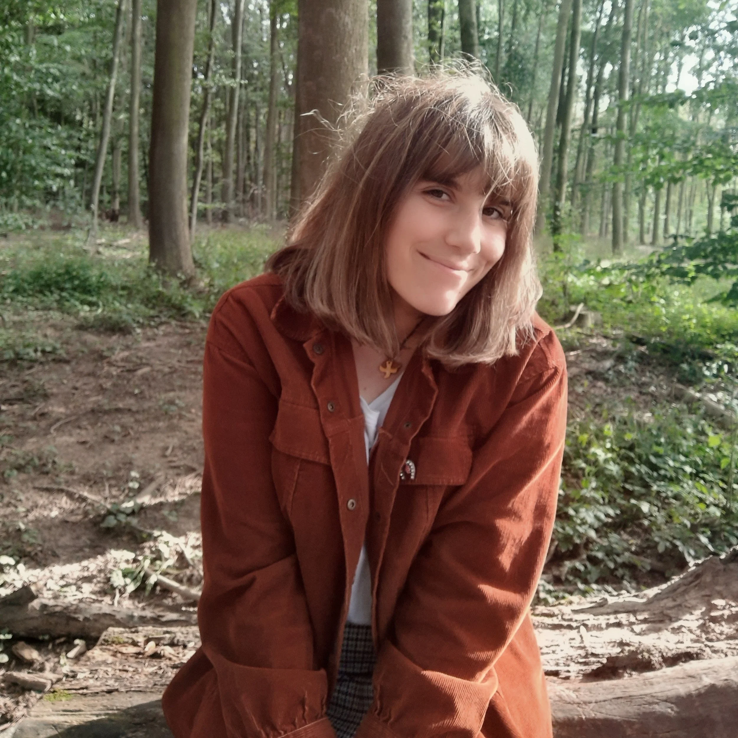 Junge Frau mit braunen Haaren im Wald, trägt braune Jacke und weißes T-Shirt, lächelnd.
