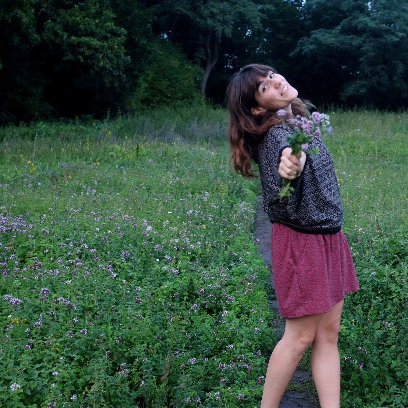 Eine junge Frau in einem roten Rock und einem schwarzen Musterpullover steht in einem blühenden Feld auf einer Waldlichtung und hält eine Blume in die Kamera.