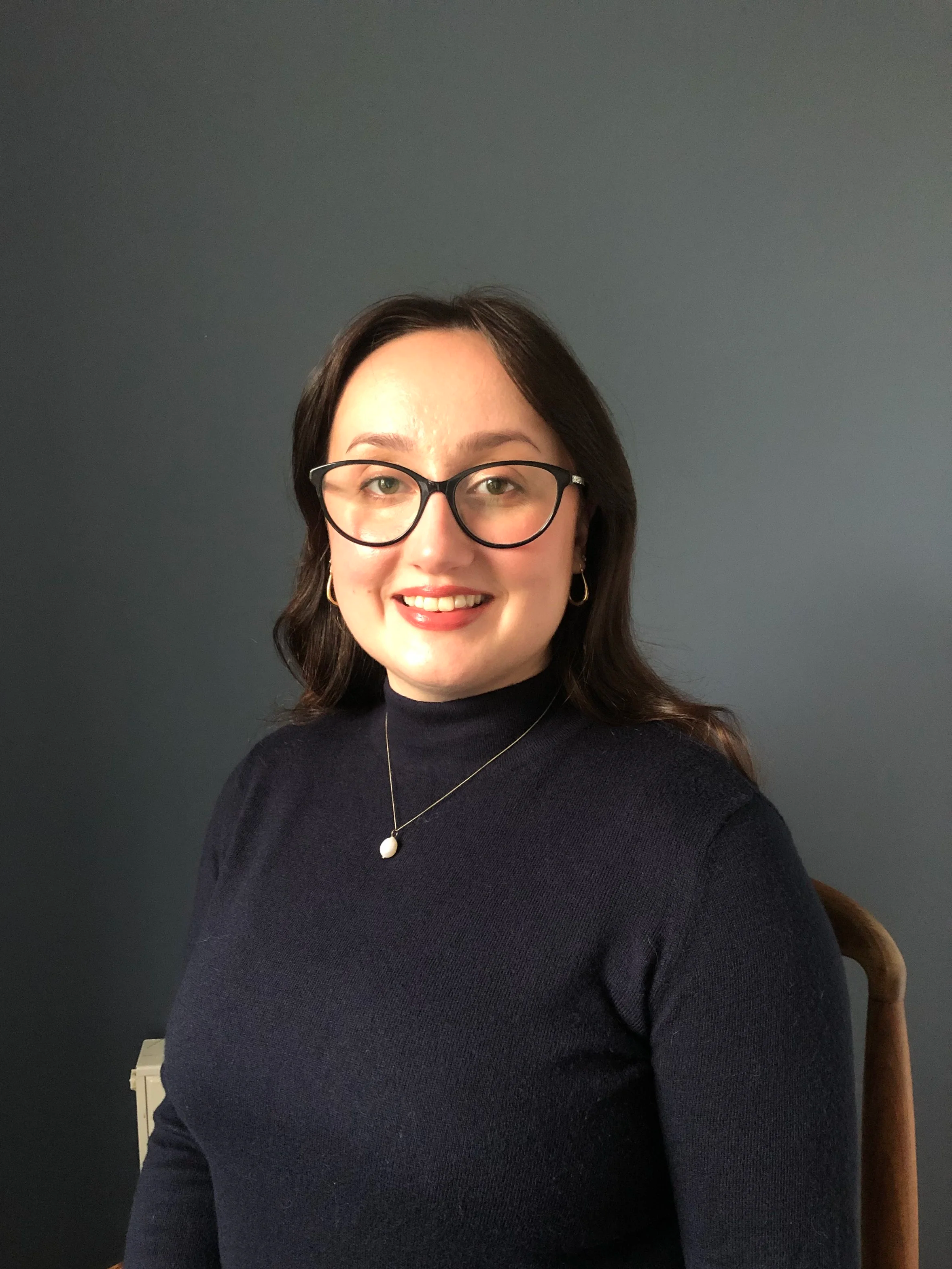 A woman with dark brown hair, glasses, and hoop earrings sitting in front of a dark gray wall, smiling at the camera, wearing a black turtleneck and a necklace with a gold pendant.