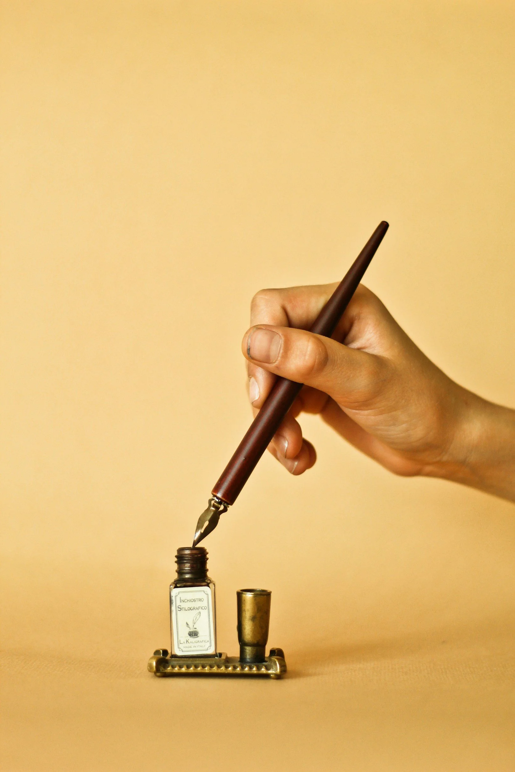 Hand holding a dip pen above an ink bottle, with an ink bottle and a brass stand on a beige background.