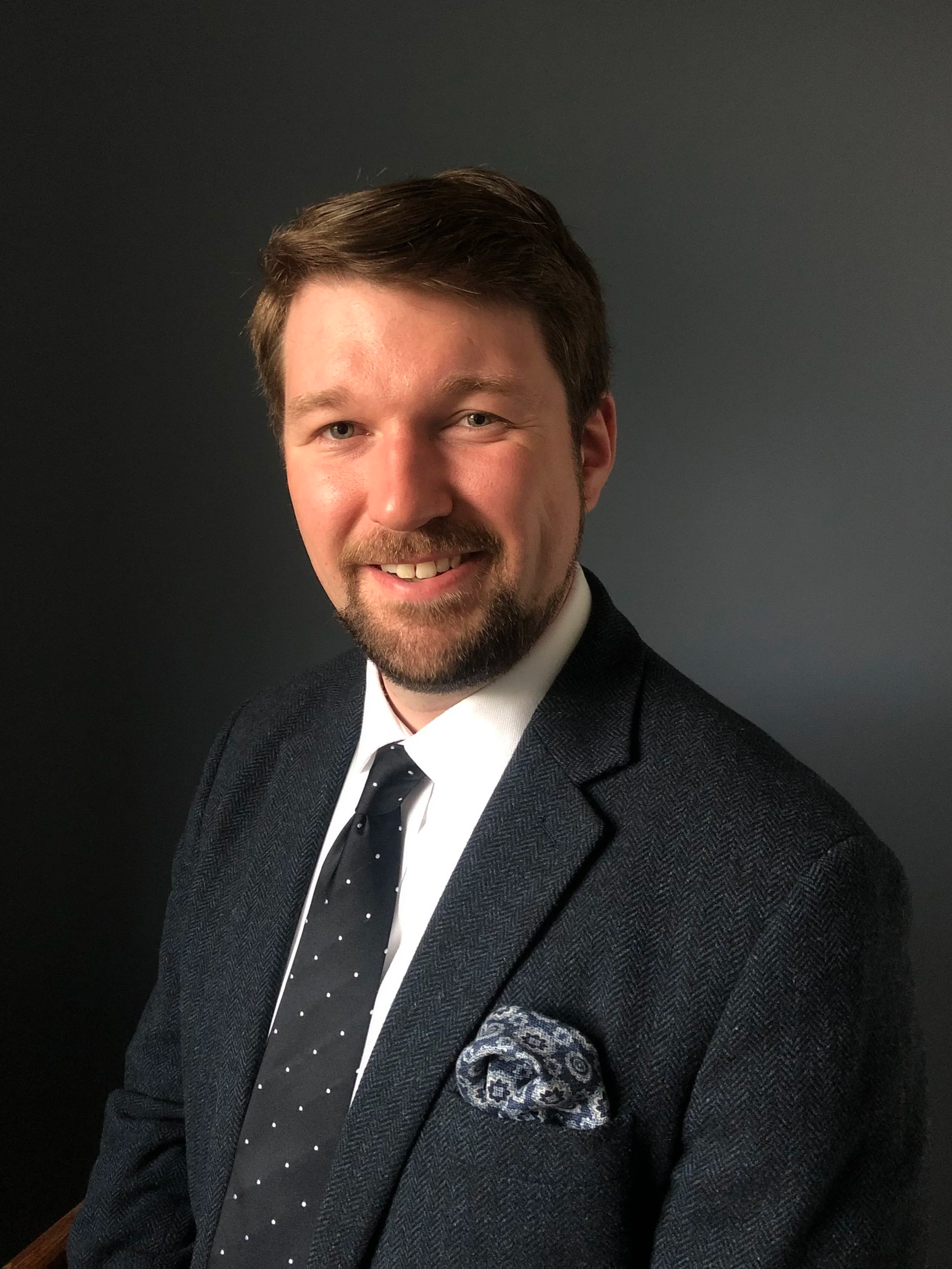 Close-up of our tutor, a smiling man with brown hair and beard, dressed in a dark suit, white shirt, and polka dot tie, against a dark background.