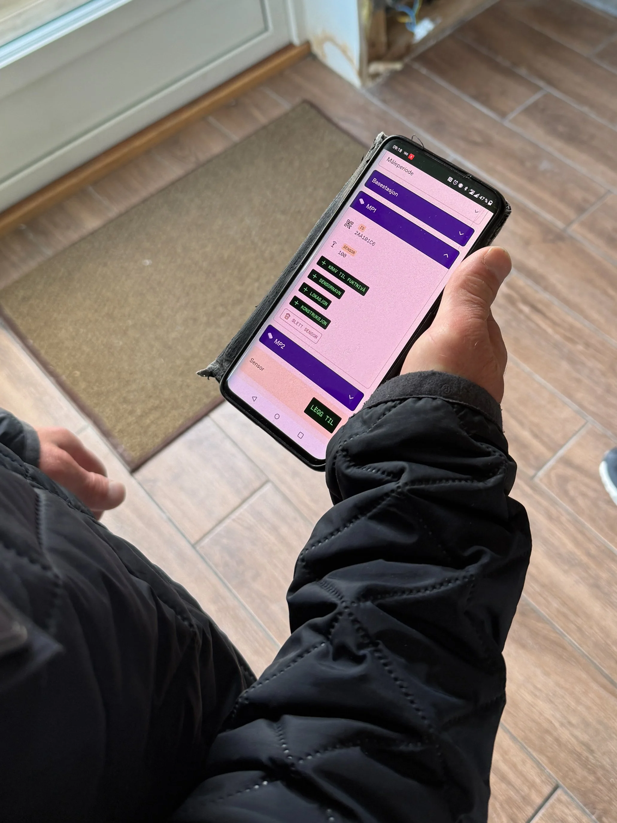 En person holder en smarttelefon som viser et oppsett for et sensorbasert system innendørs, med knapper for å legge til komponenter.