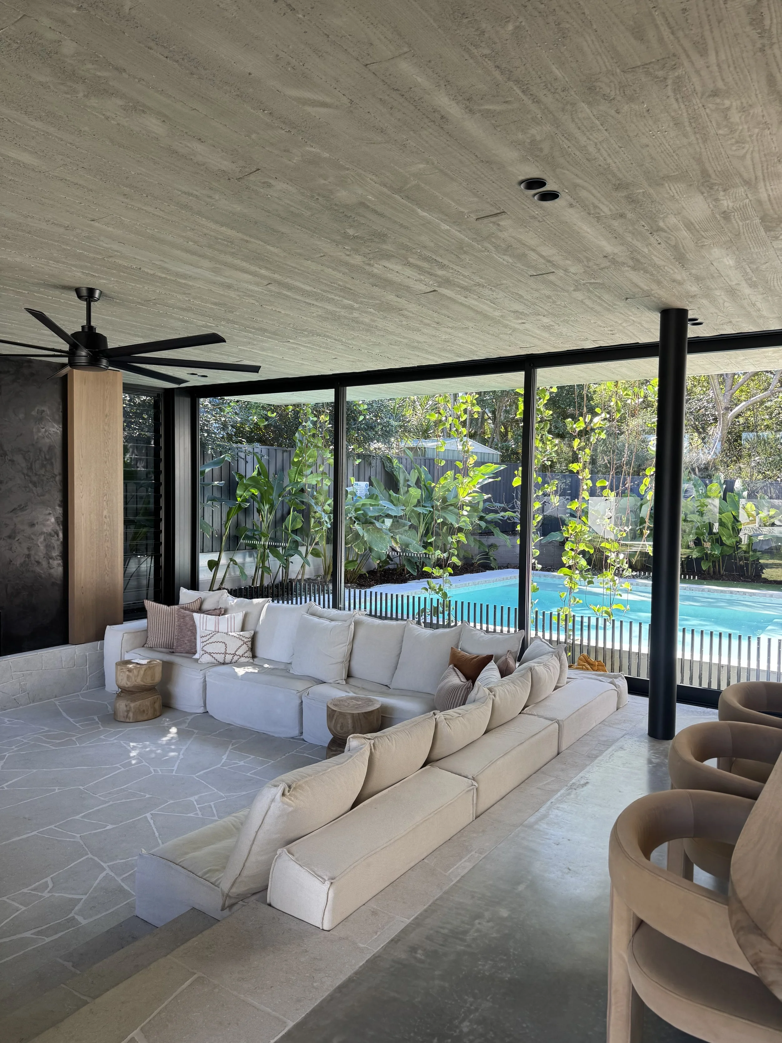 Living room with large sectional sofa, wooden side tables, ceiling fan, and a wall of floor-to-ceiling windows revealing trees and a swimming pool outside.