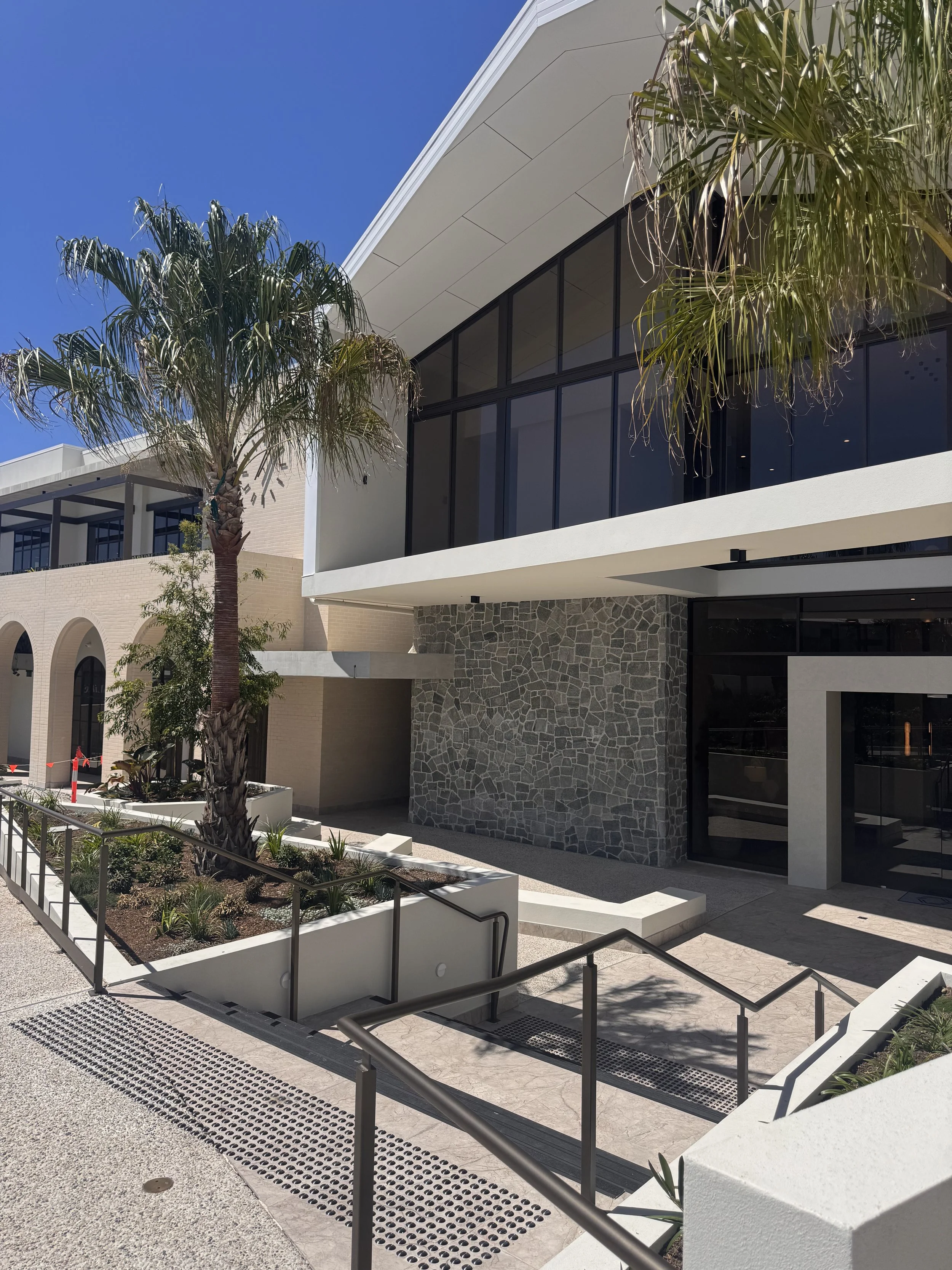 Modern commercial building with large glass windows, stone wall accents, and palm trees in planters outside. Clear blue sky.