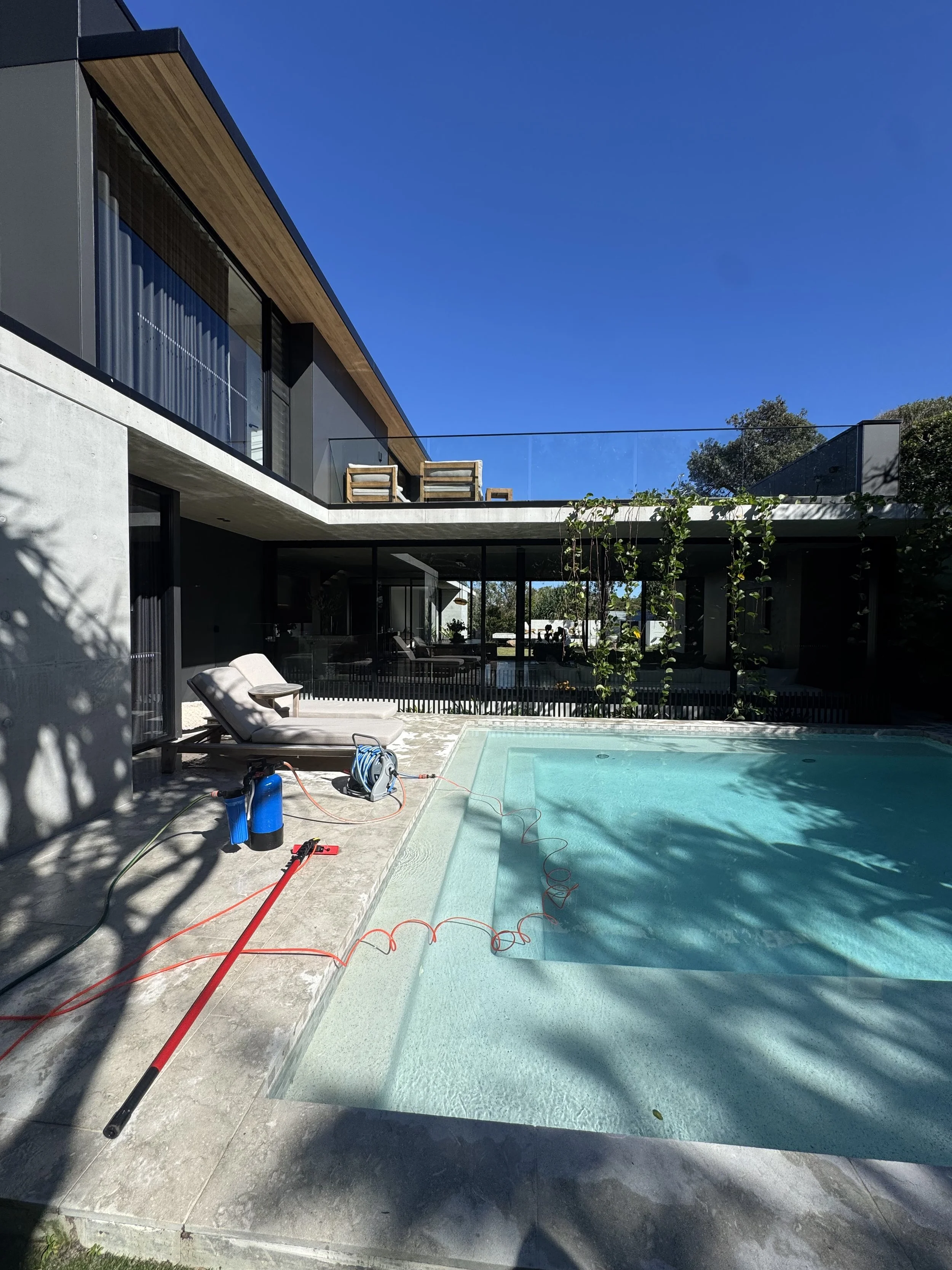 Modern house with a swimming pool in the backyard and lounge chairs, clear blue sky, and trees in the background.