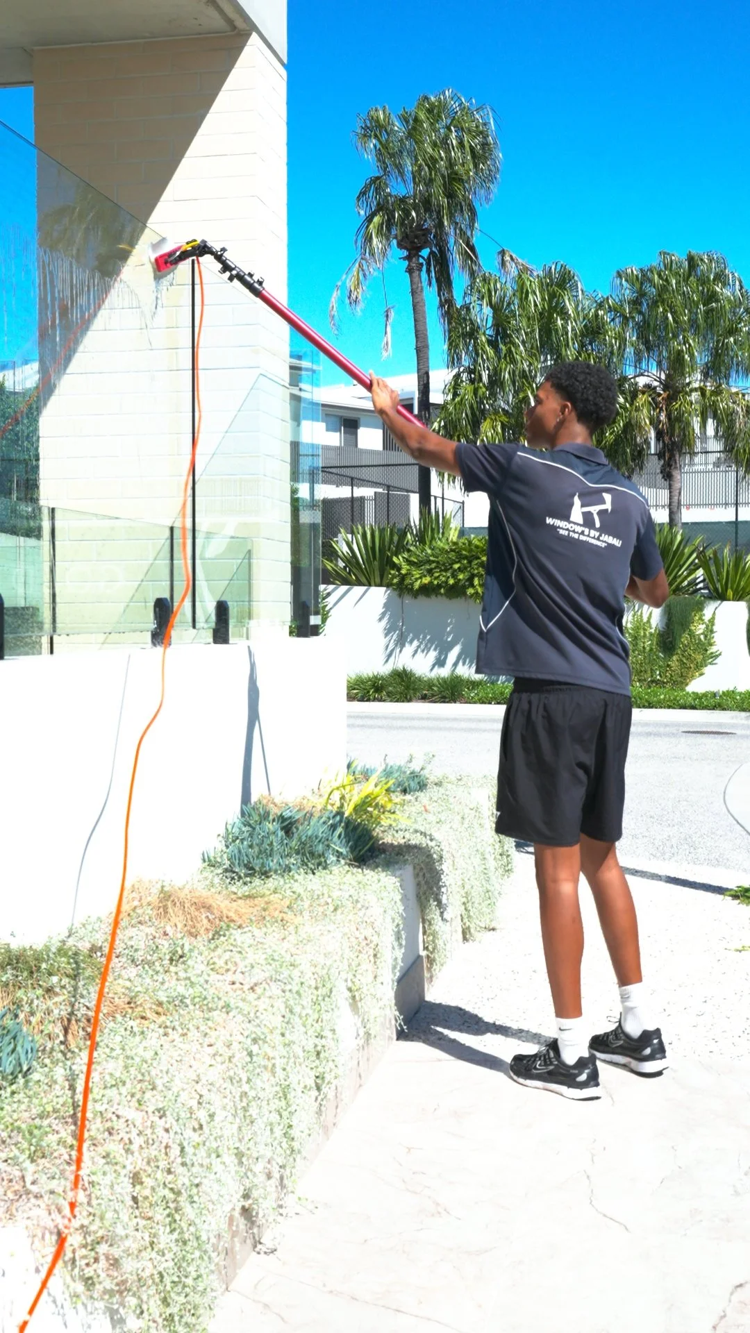 A person cleaning a glass wall with a long-handled squeegee on a sunny day, with palm trees and modern buildings in the background.
