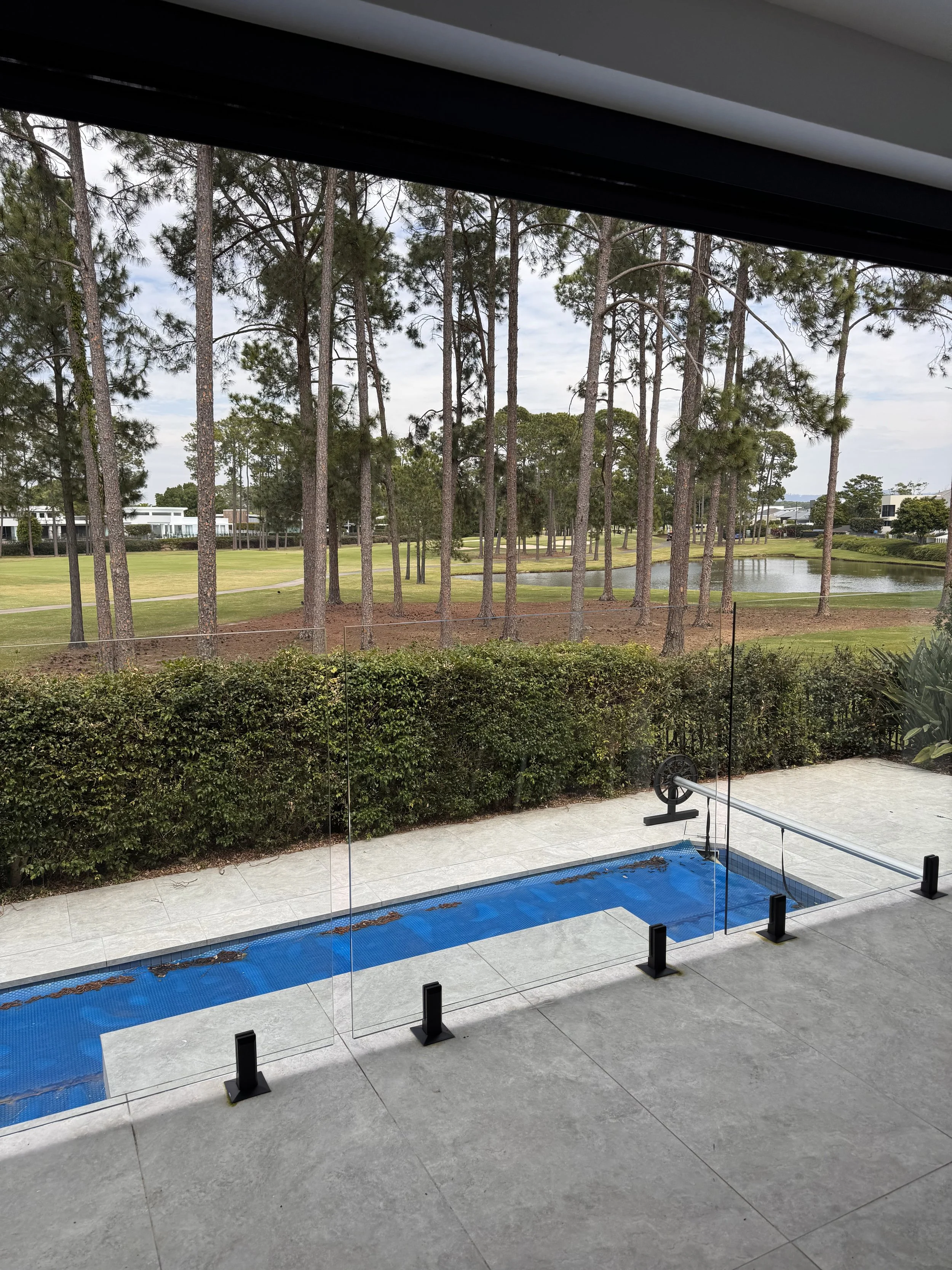 Interior view of a modern glass balcony or patio with a transparent glass railing, overlooking a landscape with a grassy area, tall trees, a pond, and contemporary residential buildings in the background.