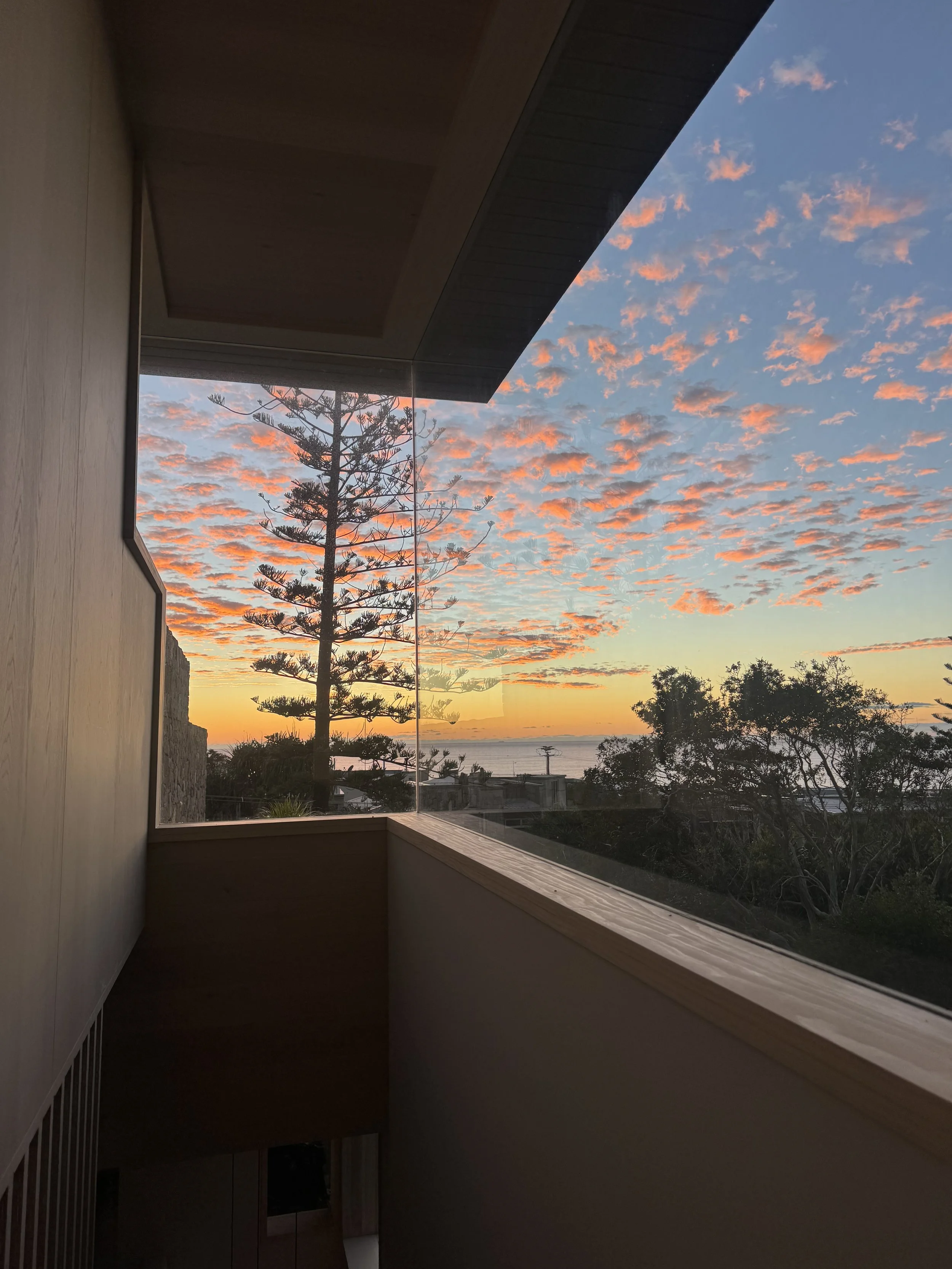 Sunset sky with pink and orange clouds seen through a large window from inside a building with wooden accents and a balcony.