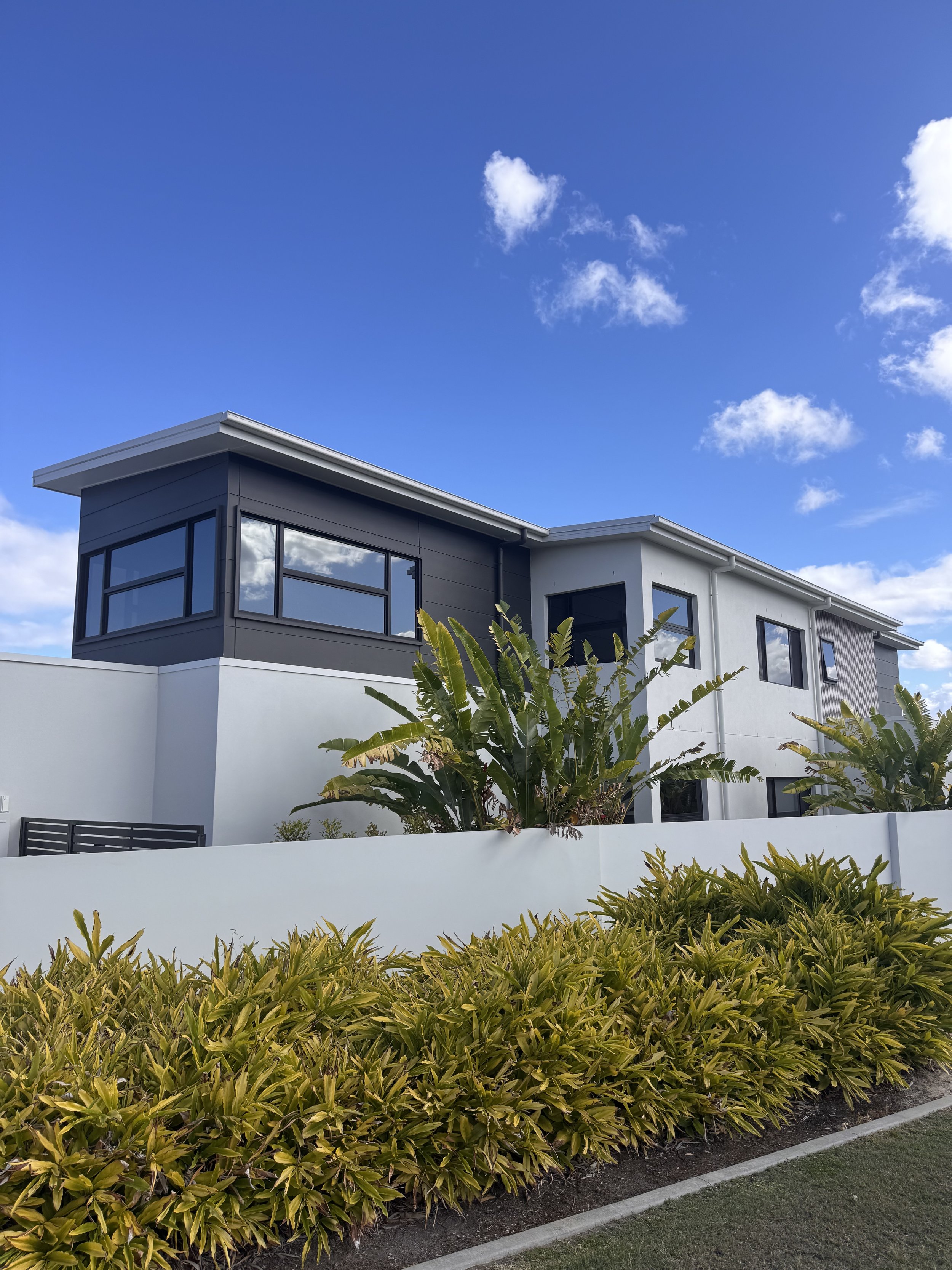 Modern two-story house with large windows, a white exterior, and a flat roof, surrounded by green and yellow shrubbery against a blue sky with a few scattered clouds.