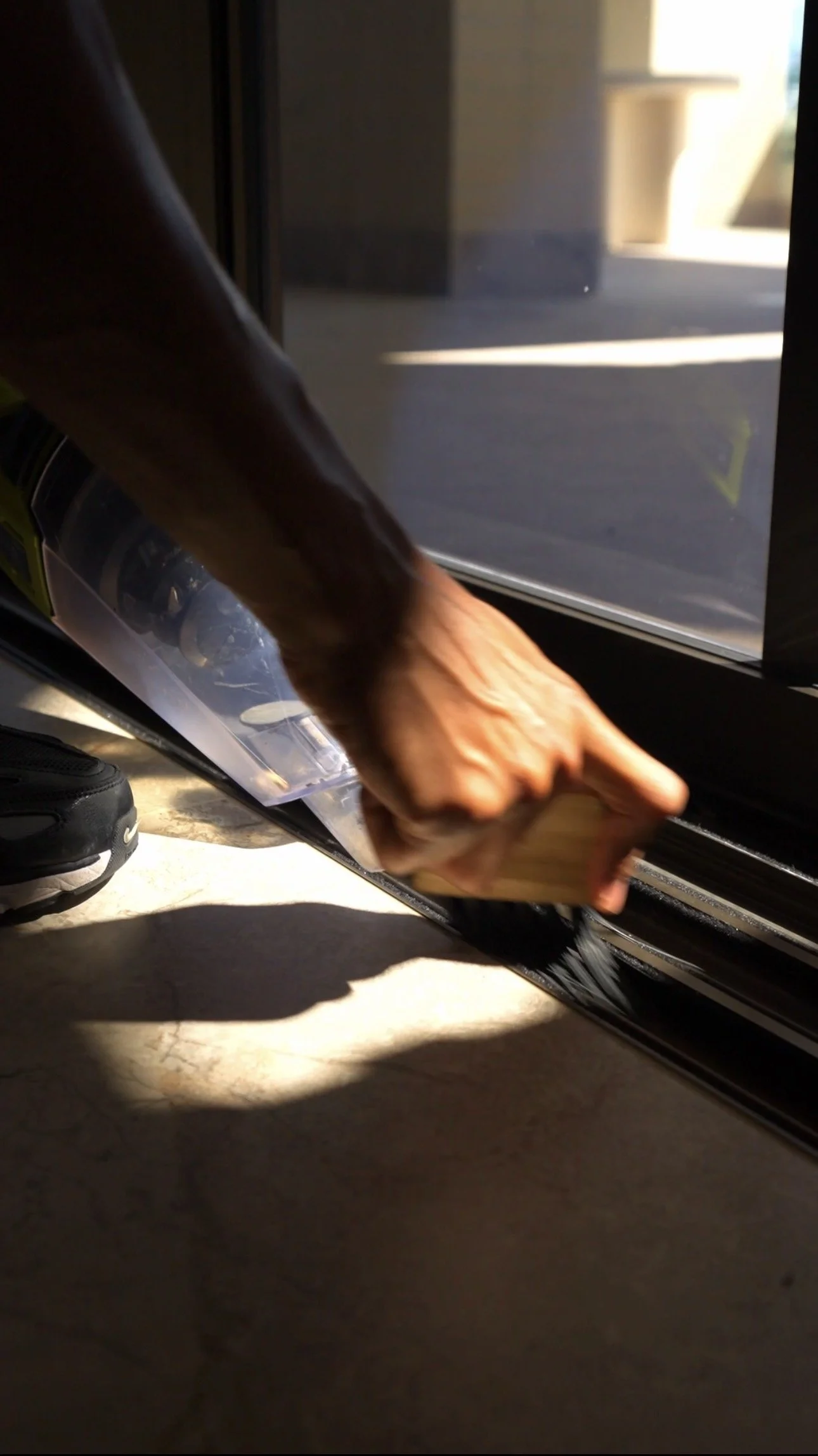 A person is cleaning a black car door with a cloth, near a glass door and shoe, with sunlight casting shadow on the floor.