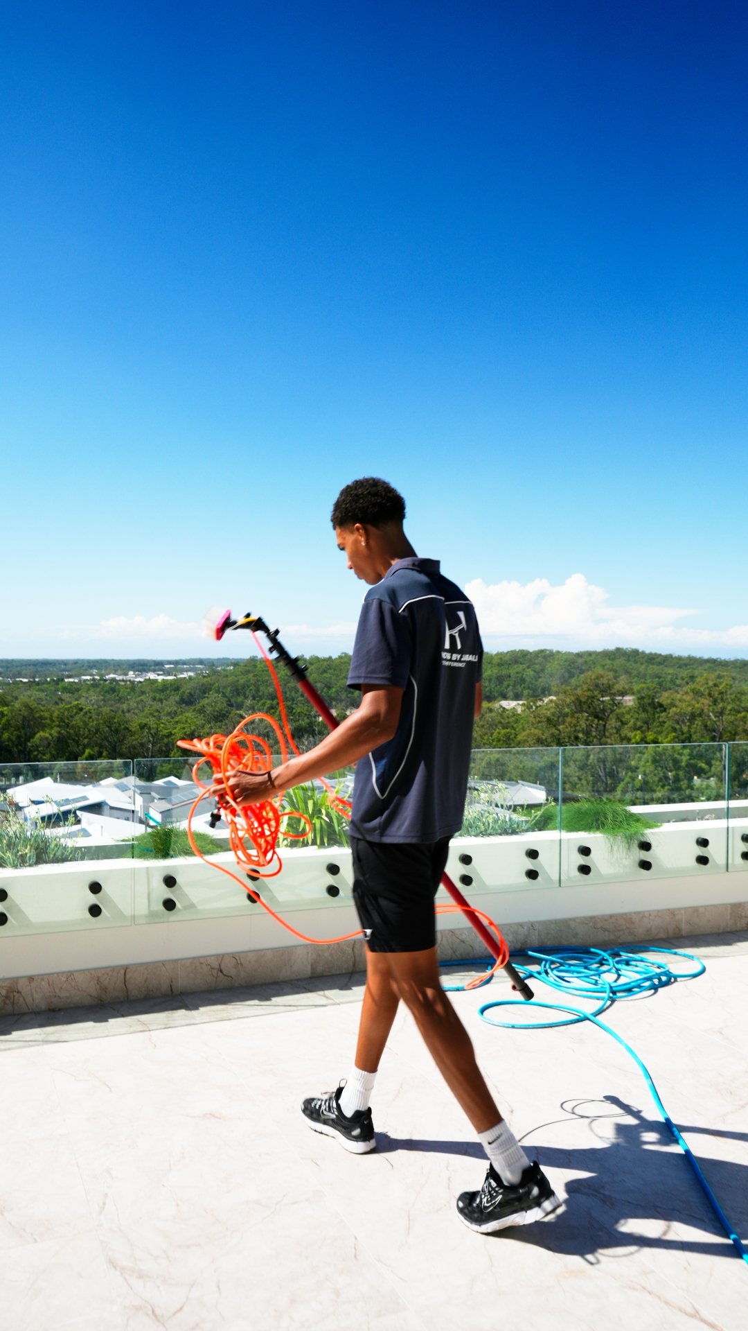 A young man walking on a rooftop terrace under a clear blue sky, holding an extension cord and an electrical saw, with a view of trees and distant buildings in the background.