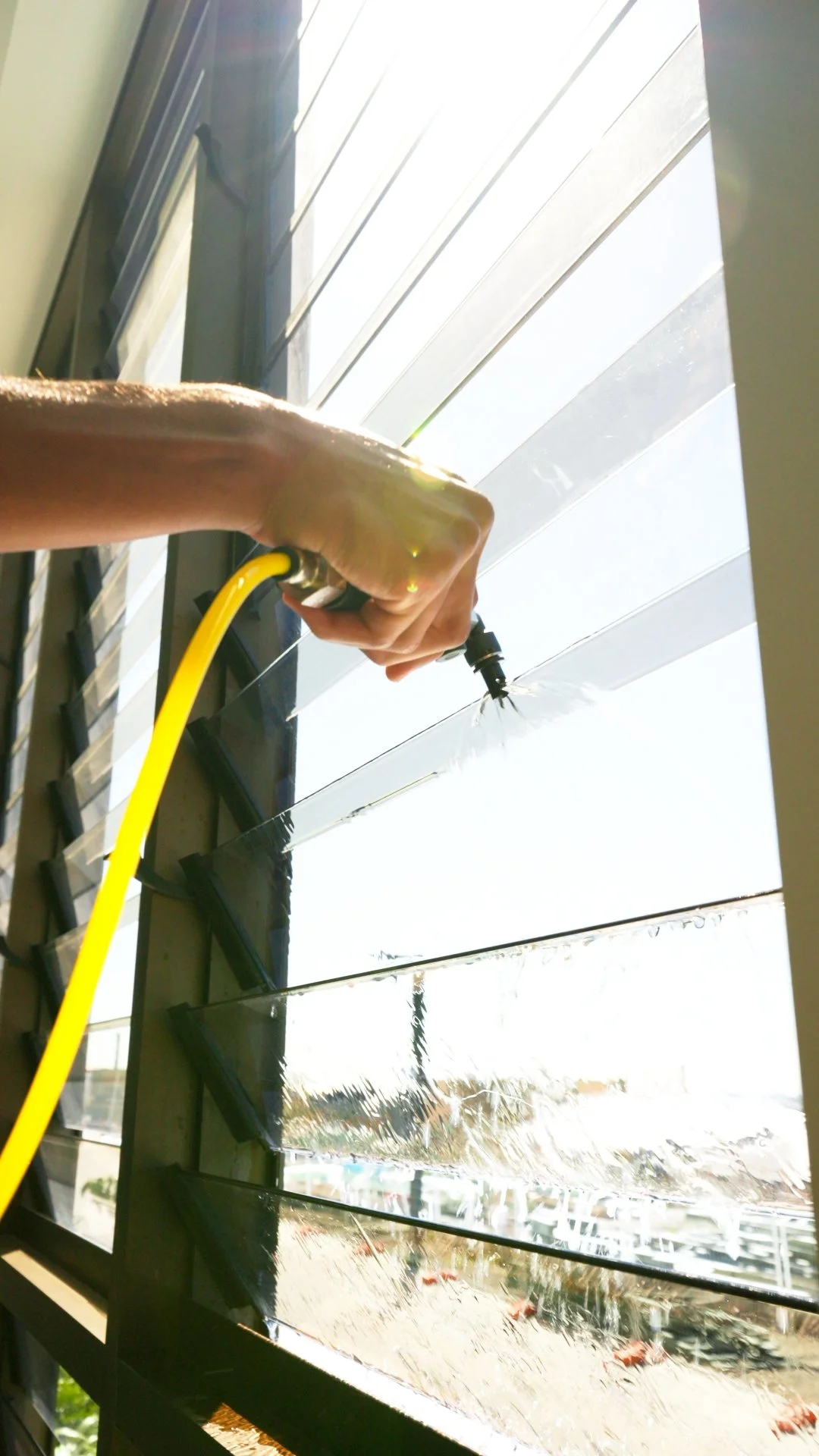 A person watering plants on a tiered window garden with a garden hose, sunlight shining through the window, and water droplets visible.