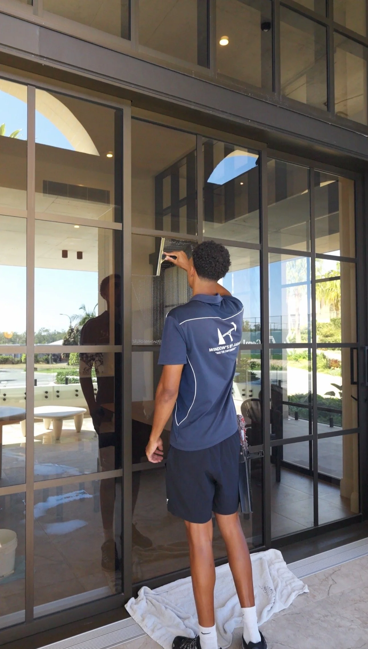 A person cleaning a glass door with a squeegee inside a modern building. The person is wearing a navy sports uniform with white and logo details, white socks, and black shoes. There is a white towel on the floor underneath them.